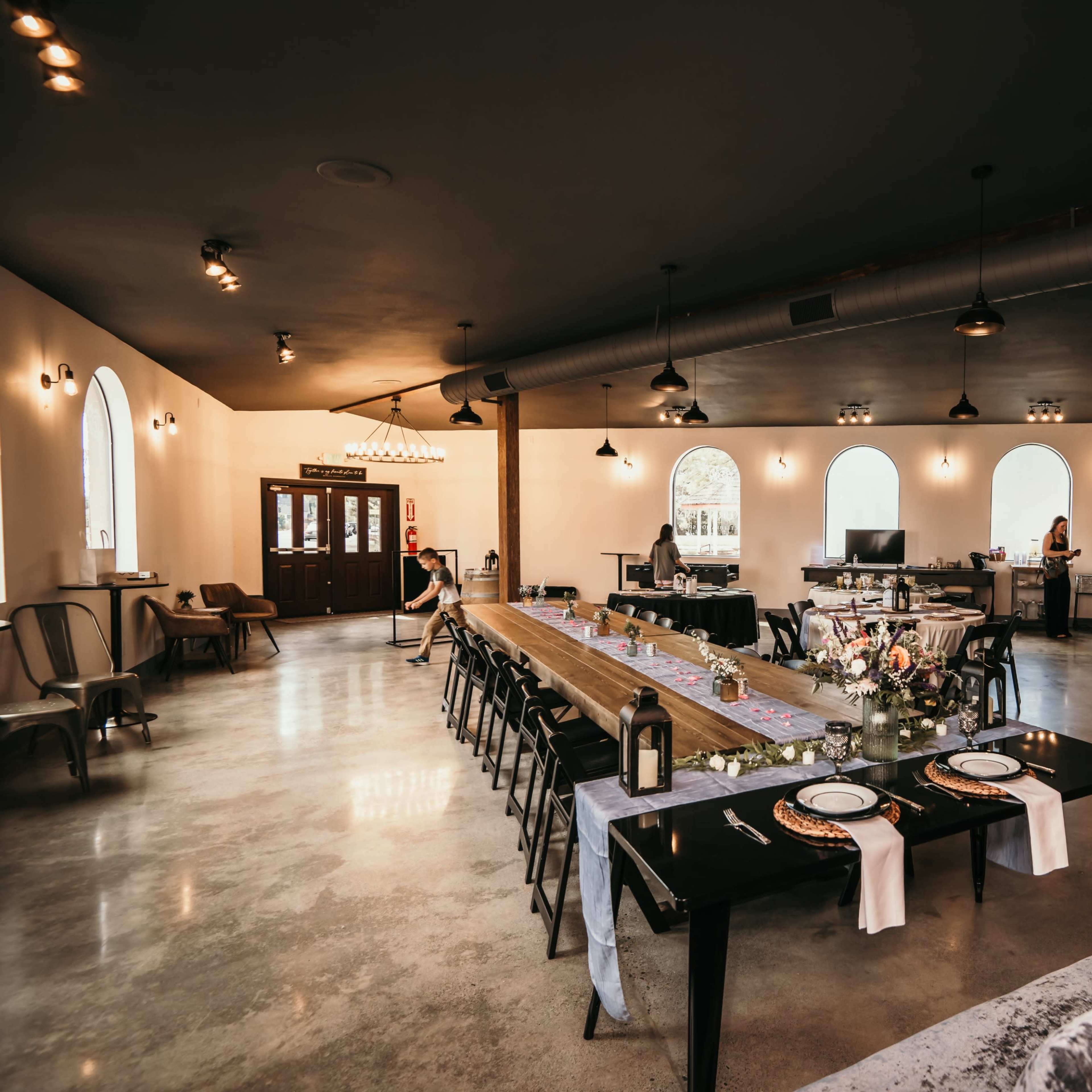A spacious dining area features a long wooden table set with plates and floral arrangements, surrounded by chairs, with large windows letting in natural light.
