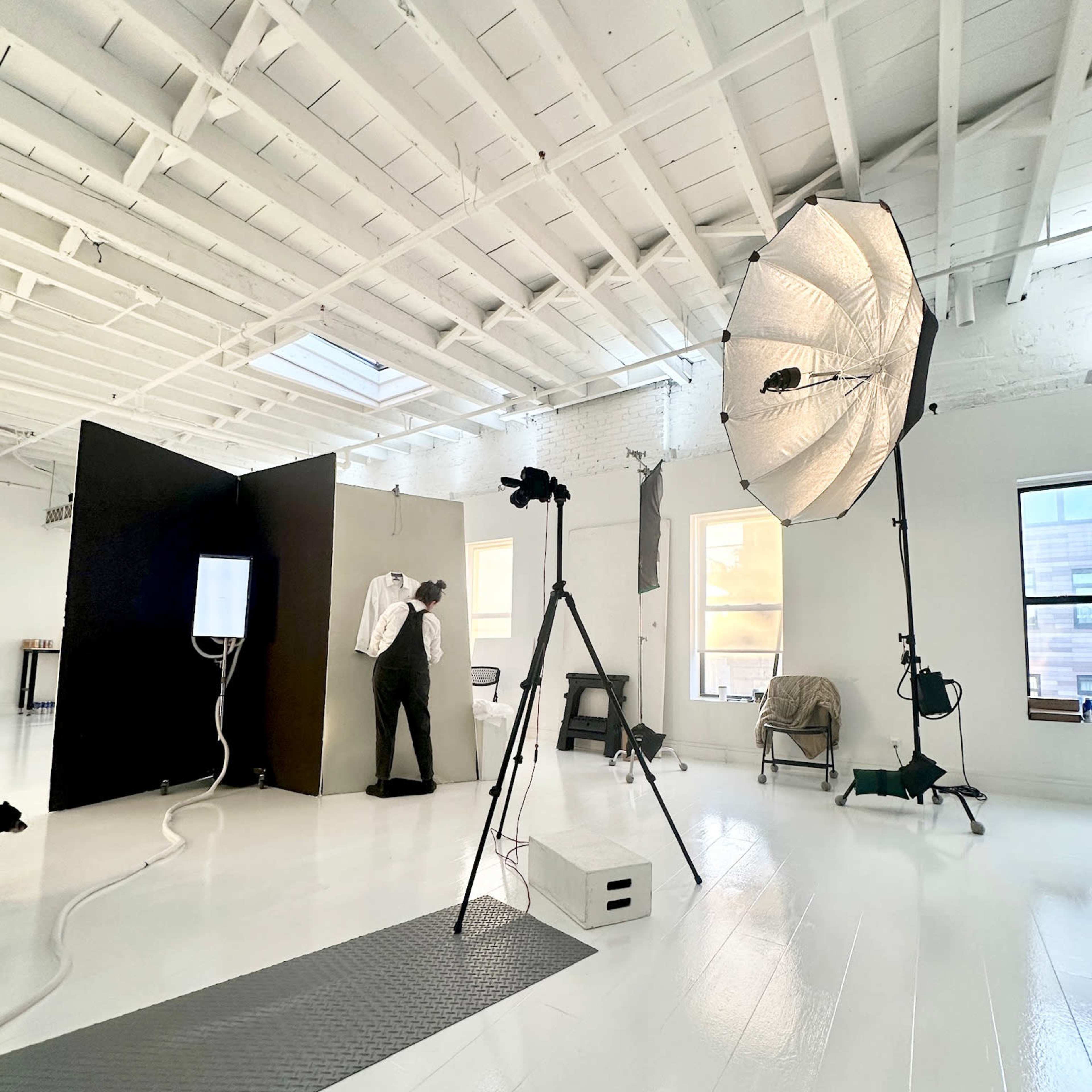A photography studio features a large softbox light, a tripod, and a setup with a model positioned against a backdrop.