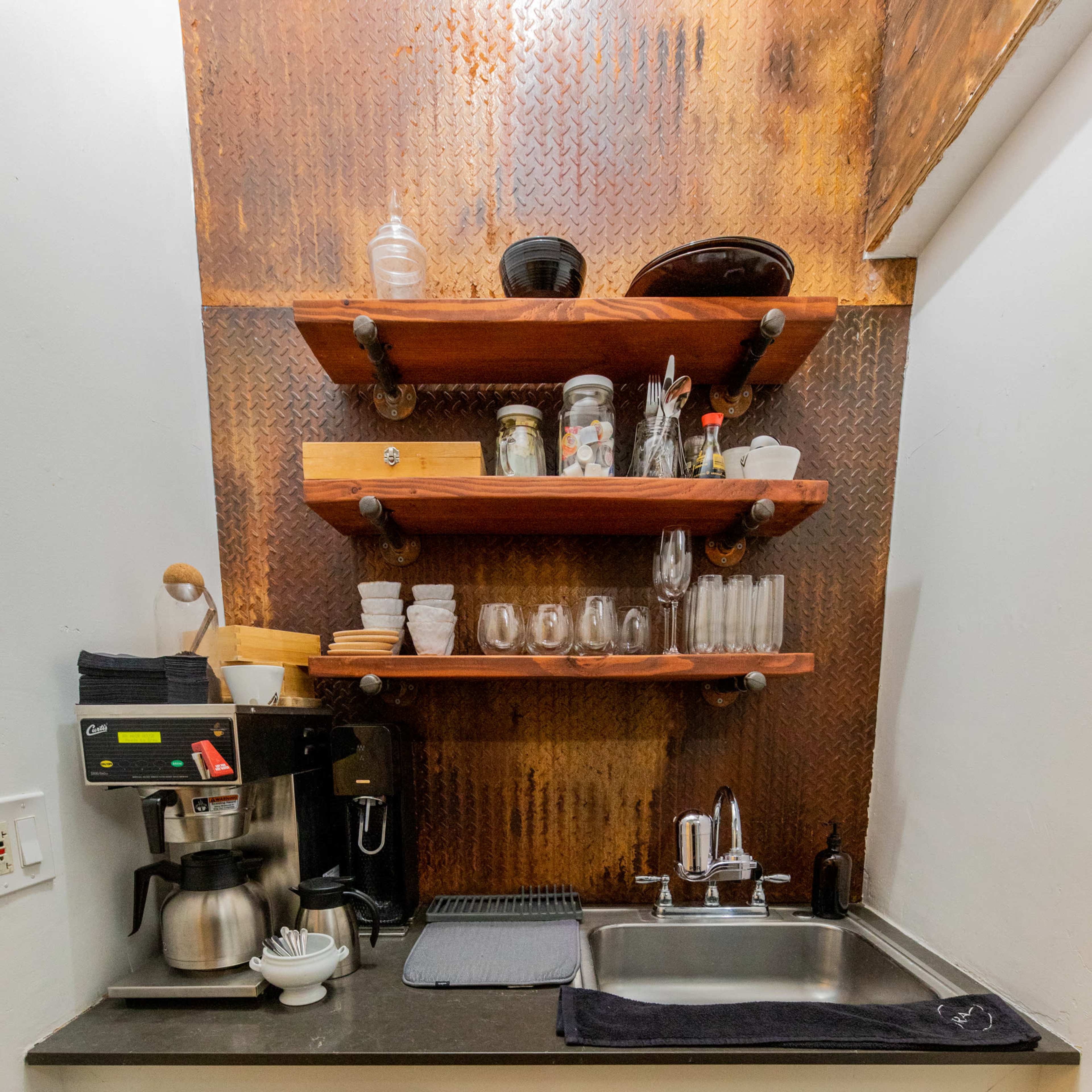 A compact kitchen area with a metal backsplash, black countertop, a coffee maker, a sink, and shelves holding various glassware and dishes.