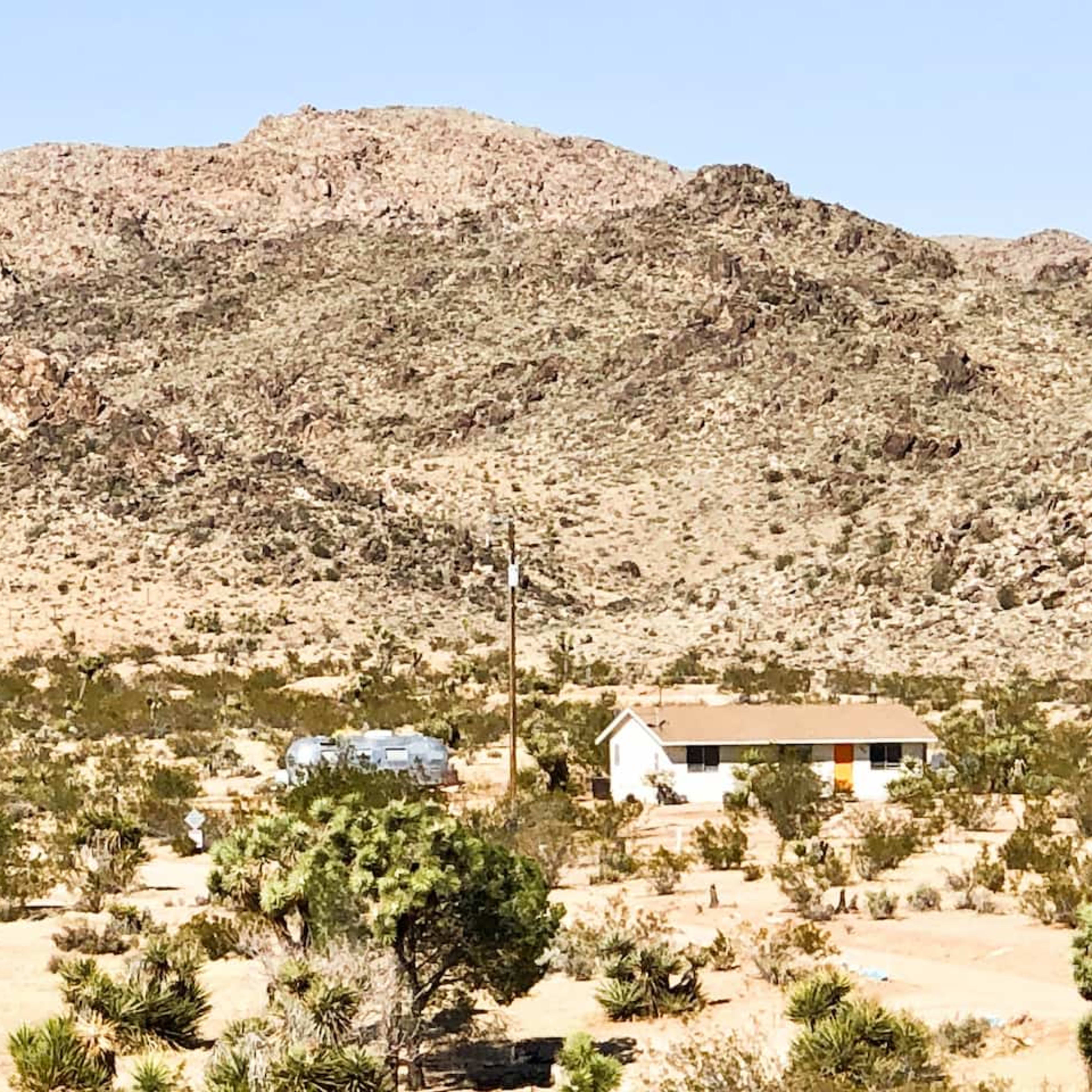 A small house situated in a desert landscape, surrounded by sparse vegetation and mountains in the background.