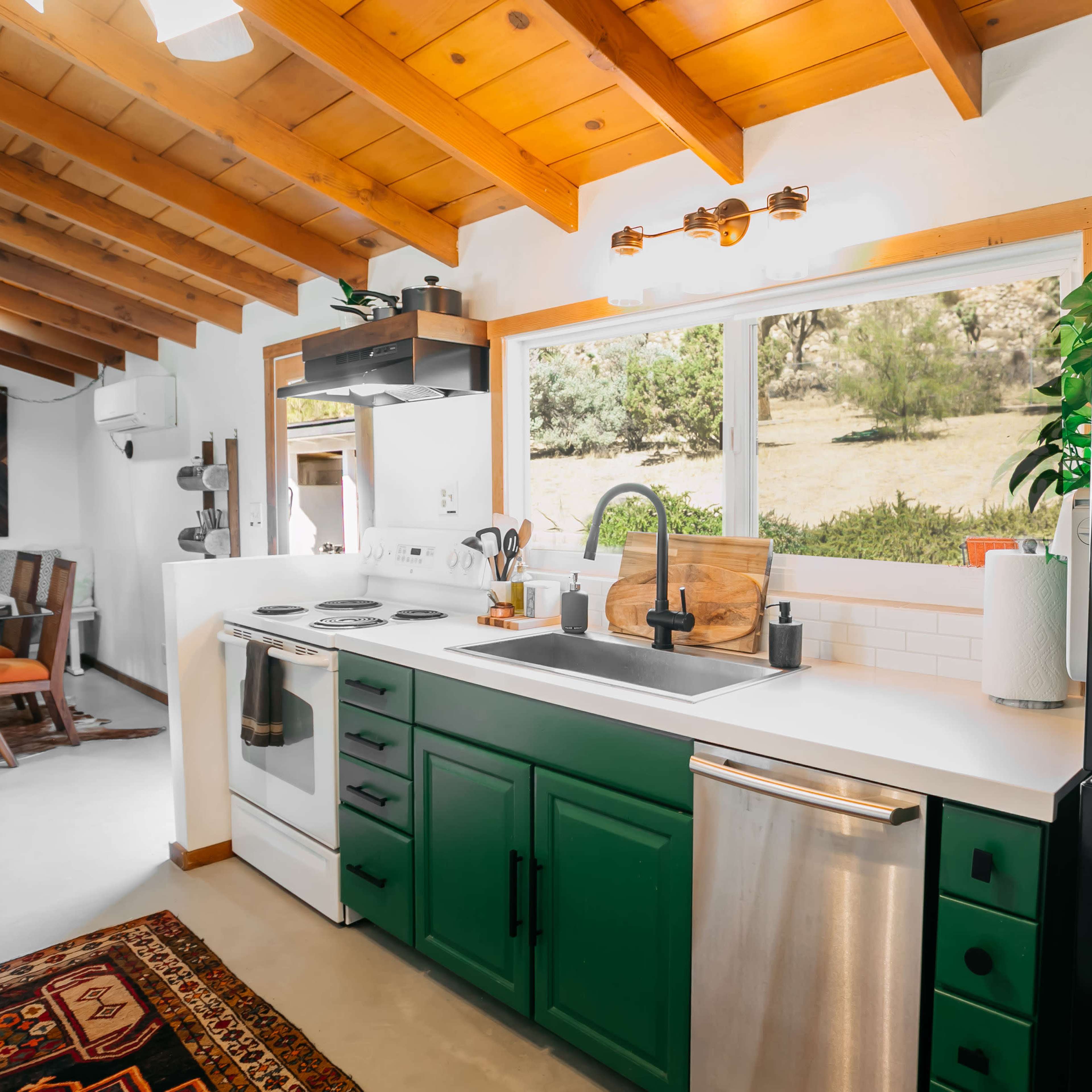 A well-designed kitchen with green cabinets, a white countertop, and a window overlooking a natural landscape.