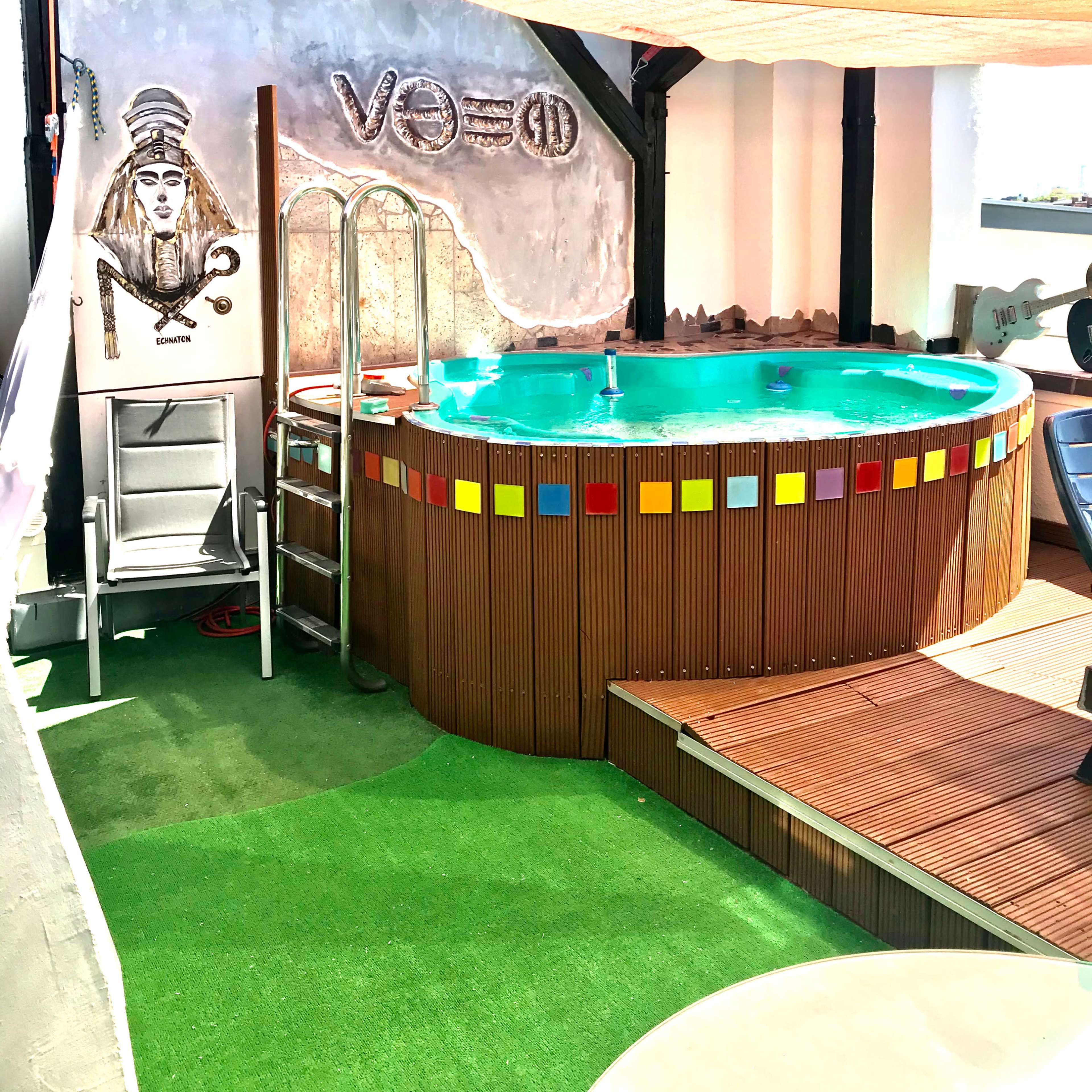 A small circular pool surrounded by a wooden deck, with a ladder on one side and a decorative wall featuring a mural.