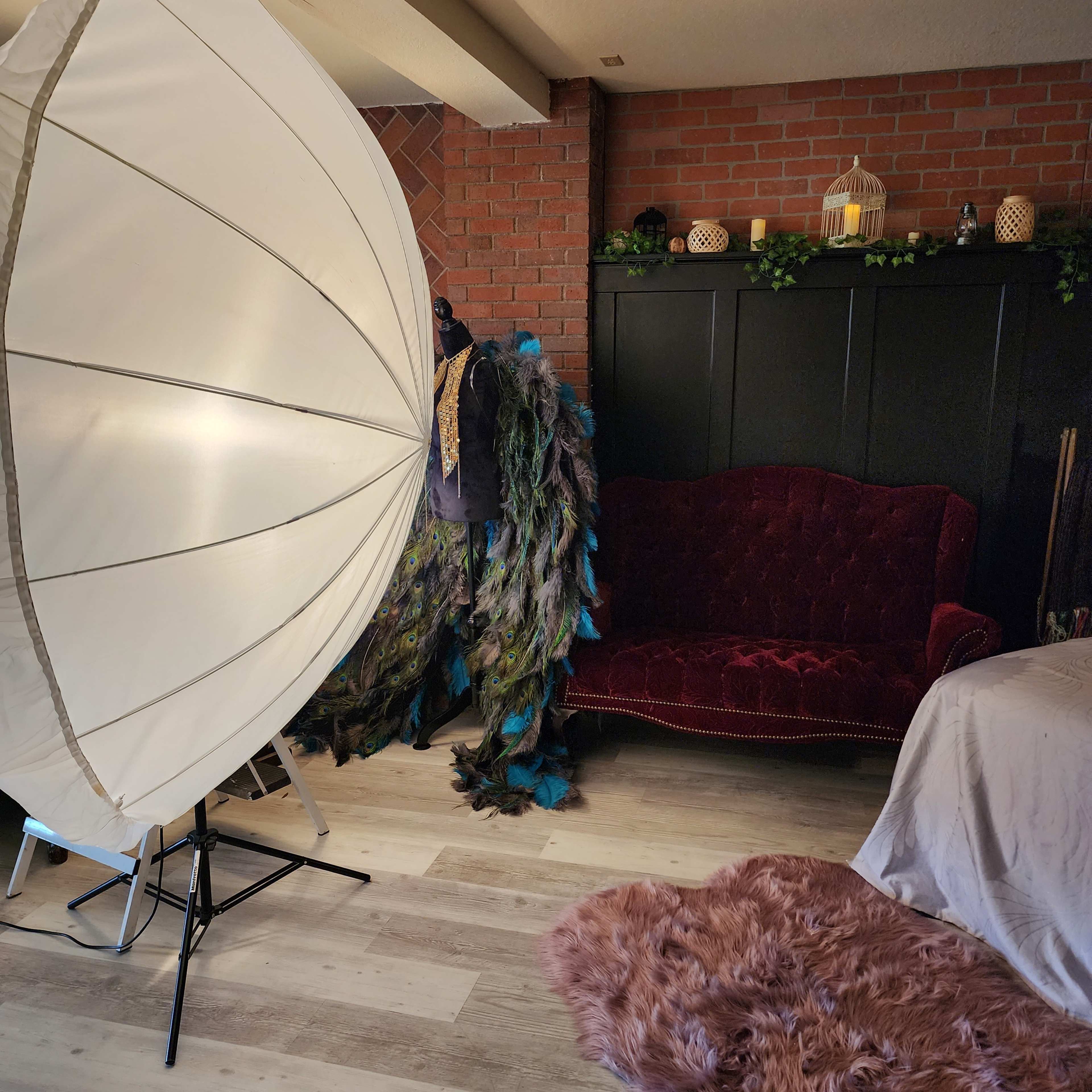 A photography studio featuring a large umbrella light, a plush red couch, and a decorative backdrop with costumes and a cozy rug.