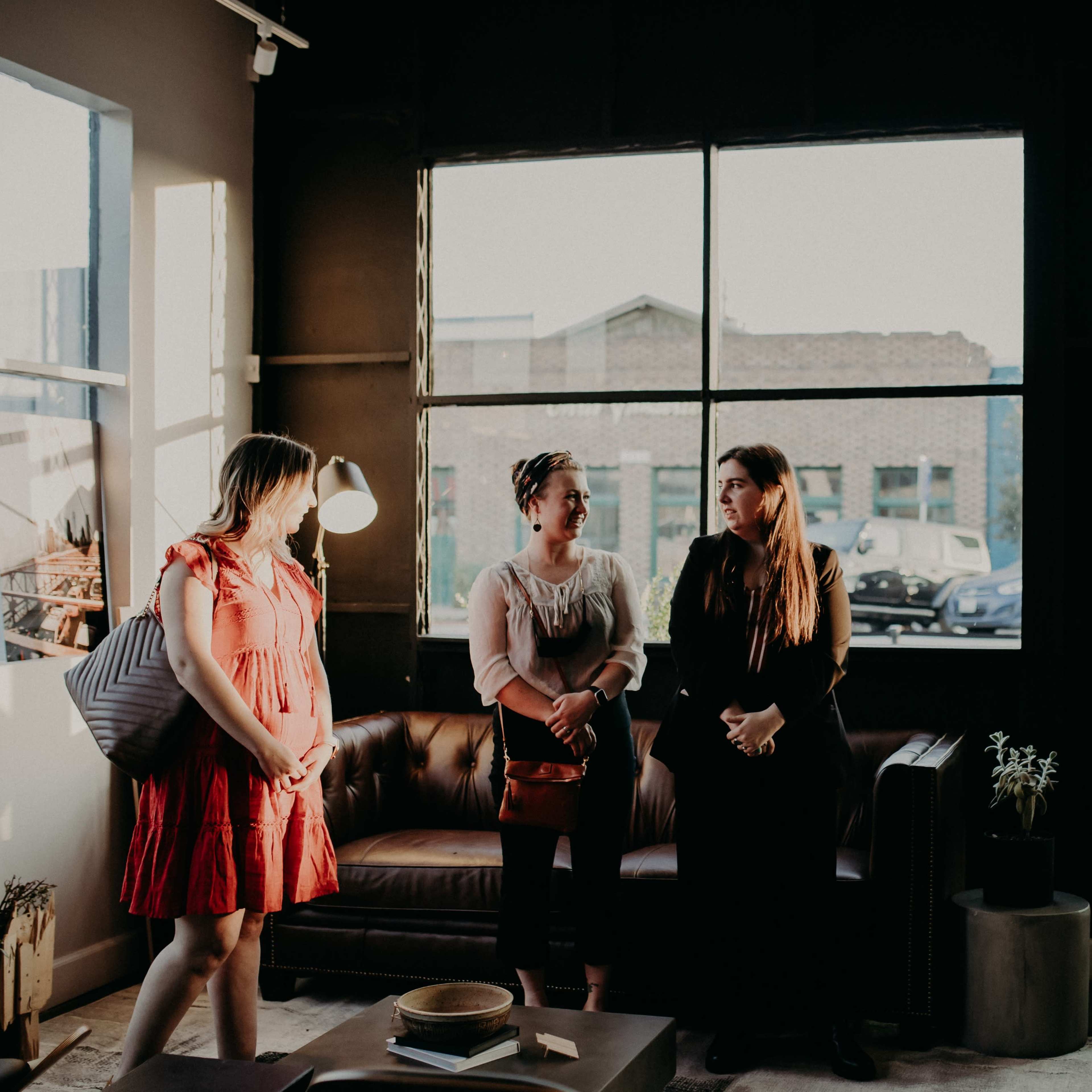 A group of women stands conversing in a modern, well-lit coffee shop with large windows.