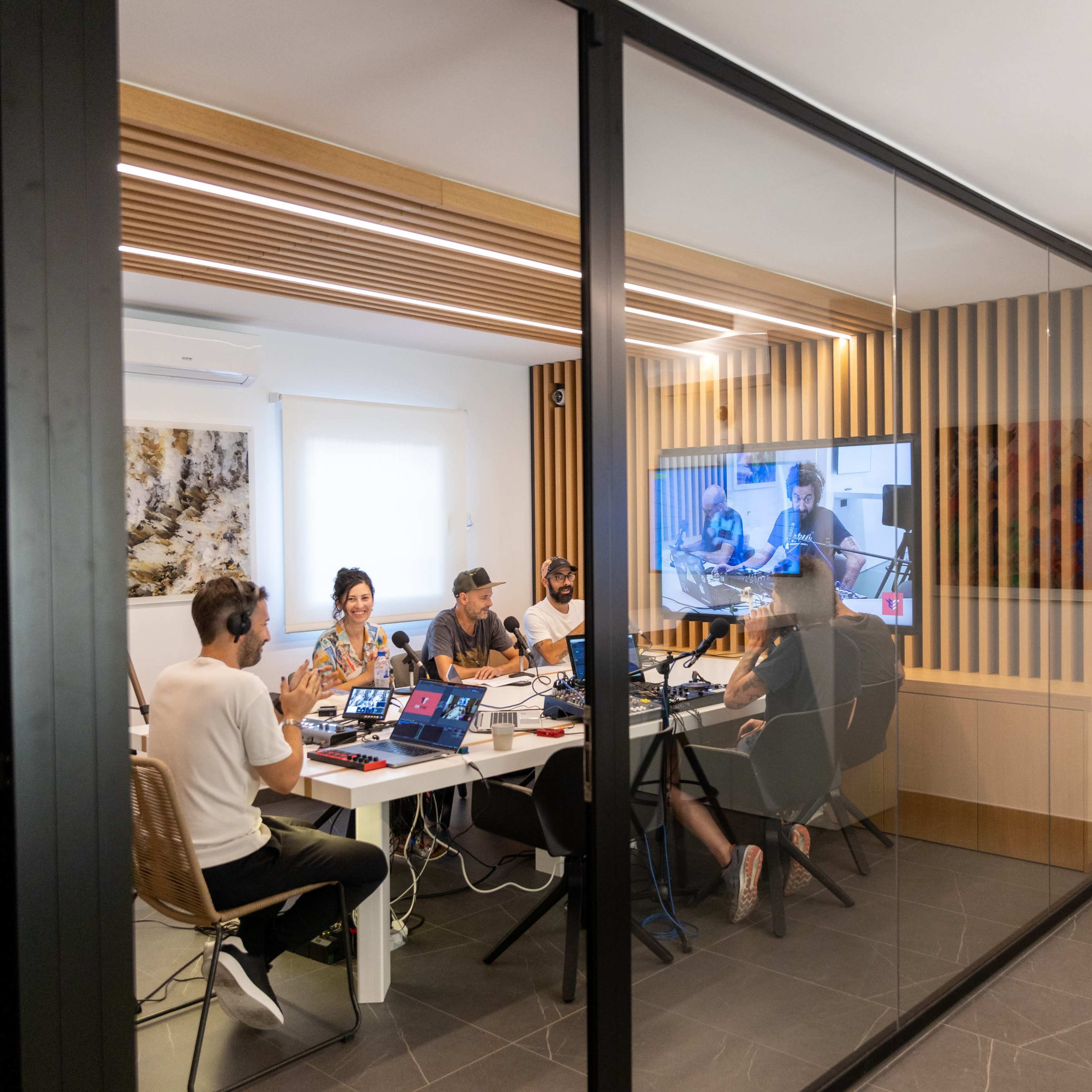 A group of five people is engaged in a video conference in a room separated by glass walls, equipped with computers and audio equipment.