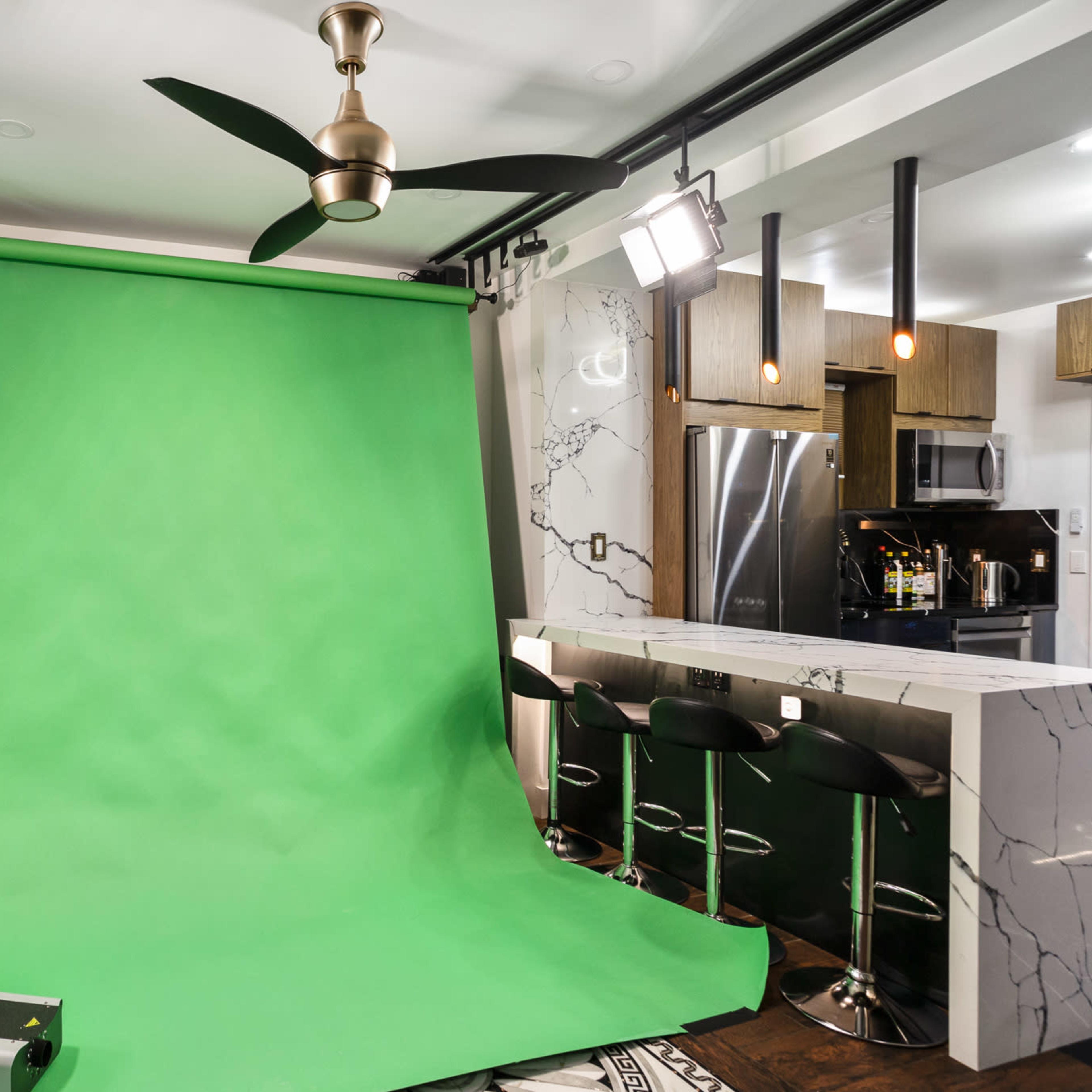 A studio setup with a green backdrop, black bar stools, and modern kitchen appliances in the background.