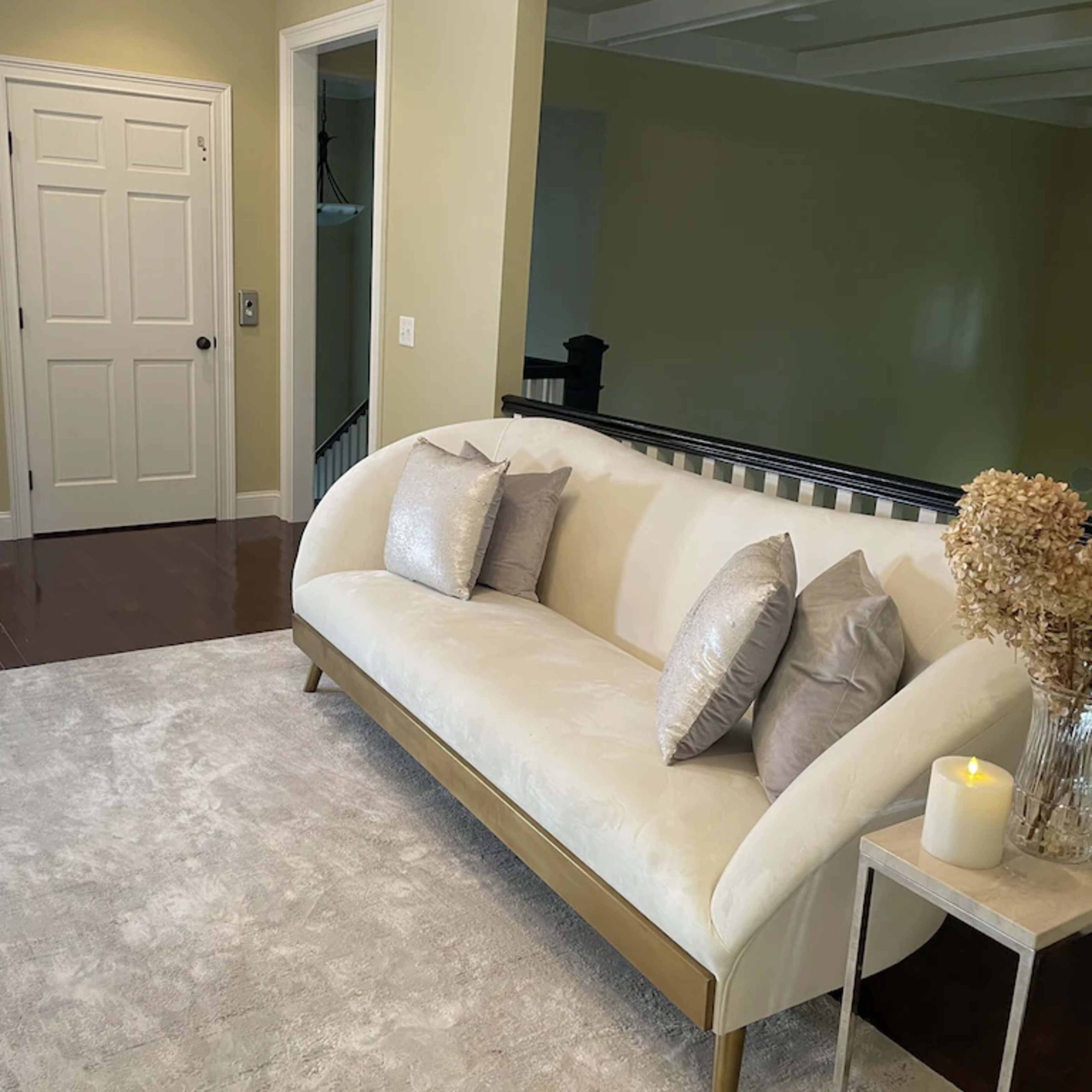 A light-colored sofa with decorative pillows placed on a plush rug in a well-lit room, with a doorway and a staircase visible in the background.