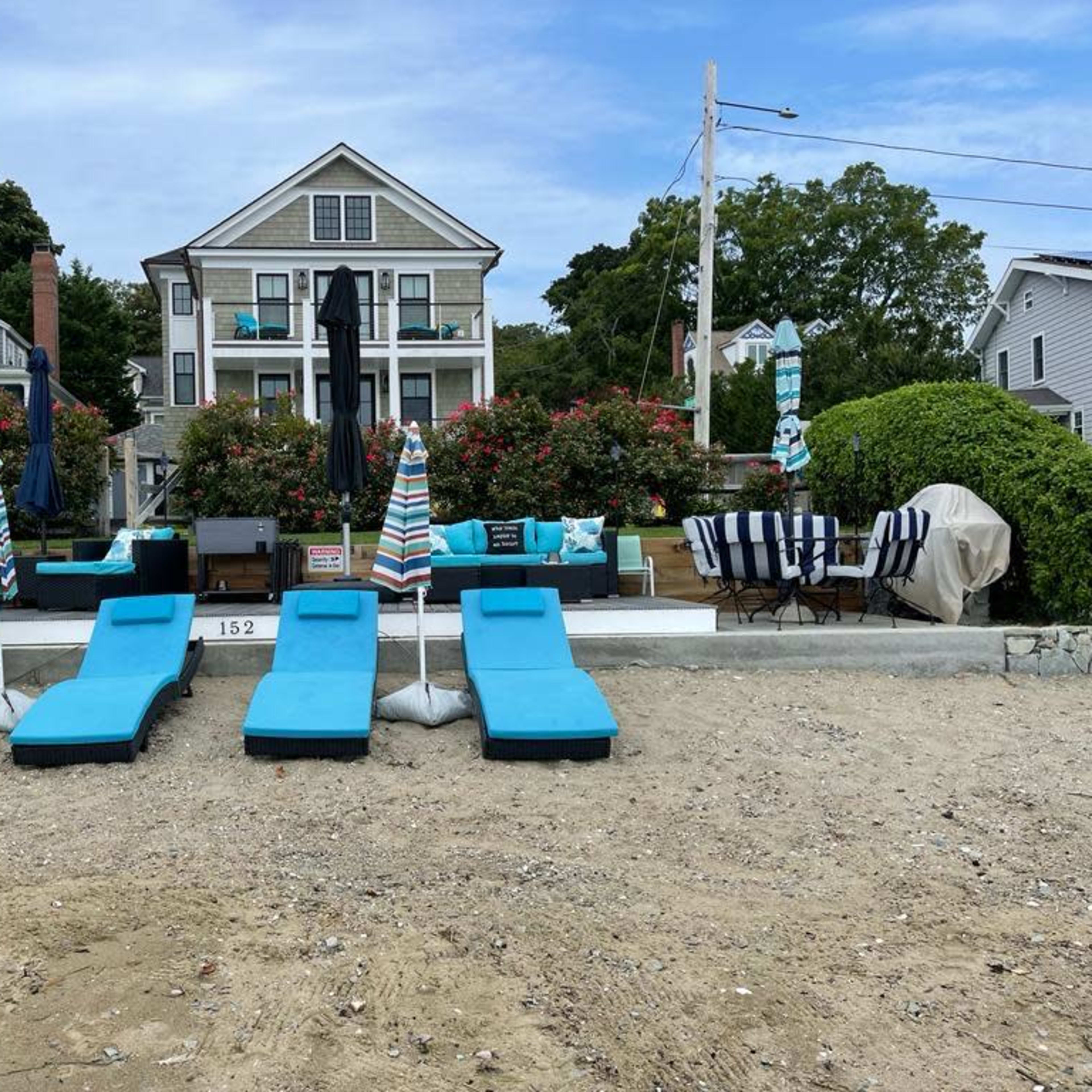 A beachside property featuring lounge chairs with umbrellas, a patio area, and a large house in the background surrounded by greenery.