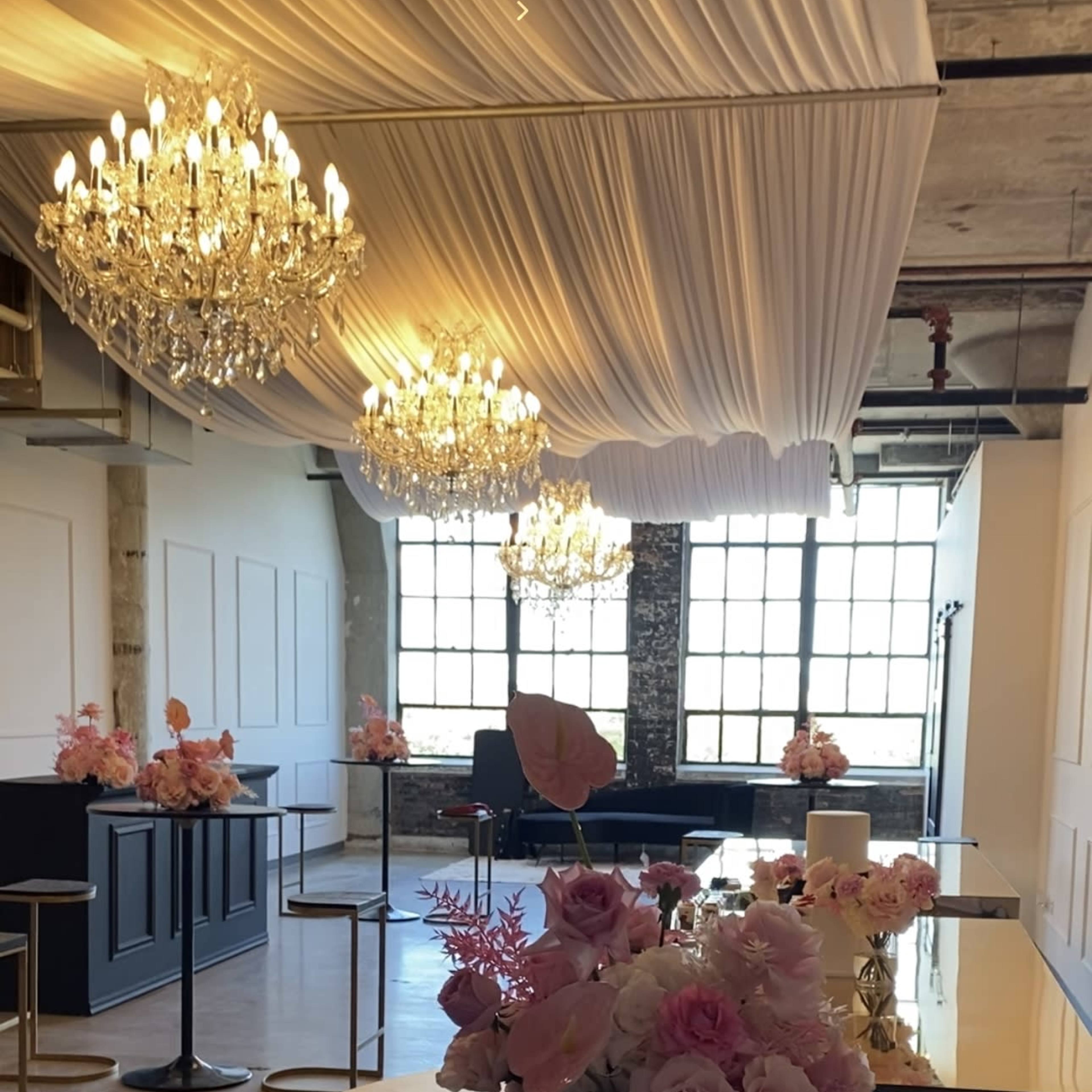 A spacious event venue with tables adorned with floral arrangements, illuminated by chandeliers hanging from a draped ceiling.