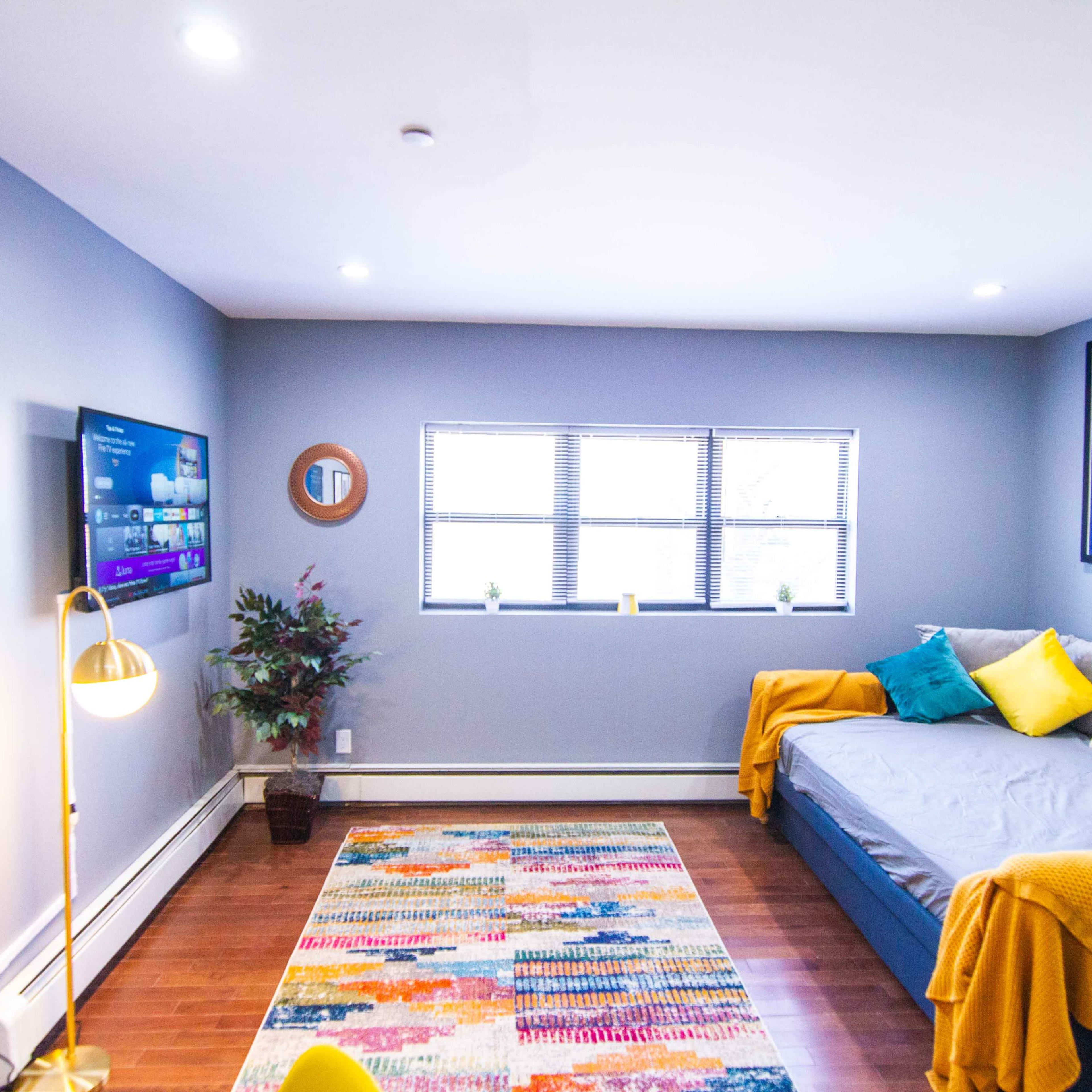 A modern living room featuring a gray wall, a television mounted on one wall, a daybed with colorful pillows, and a patterned area rug.