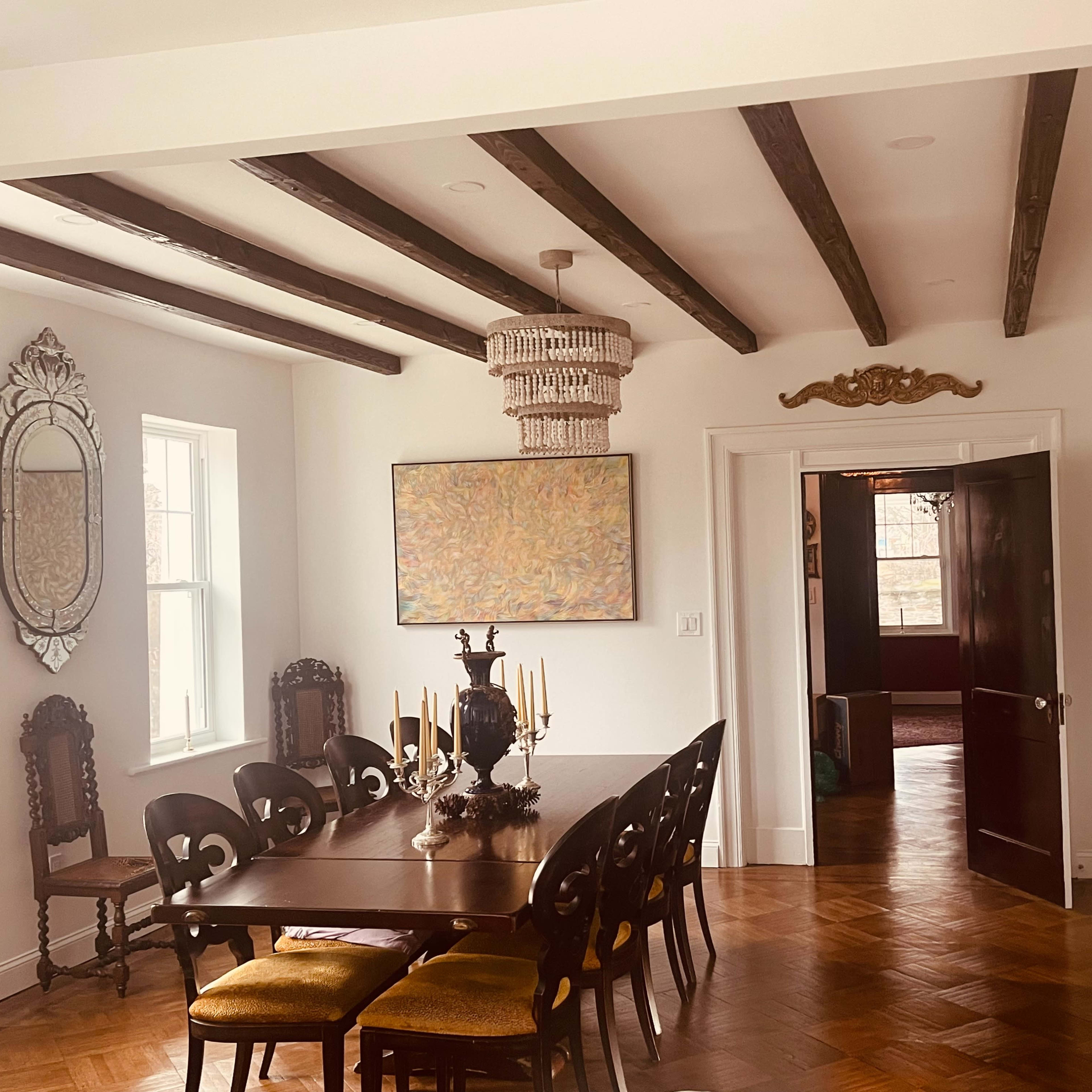 A dining room features a long wooden table surrounded by chairs, with a chandelier above and an ornate mirror on the wall.