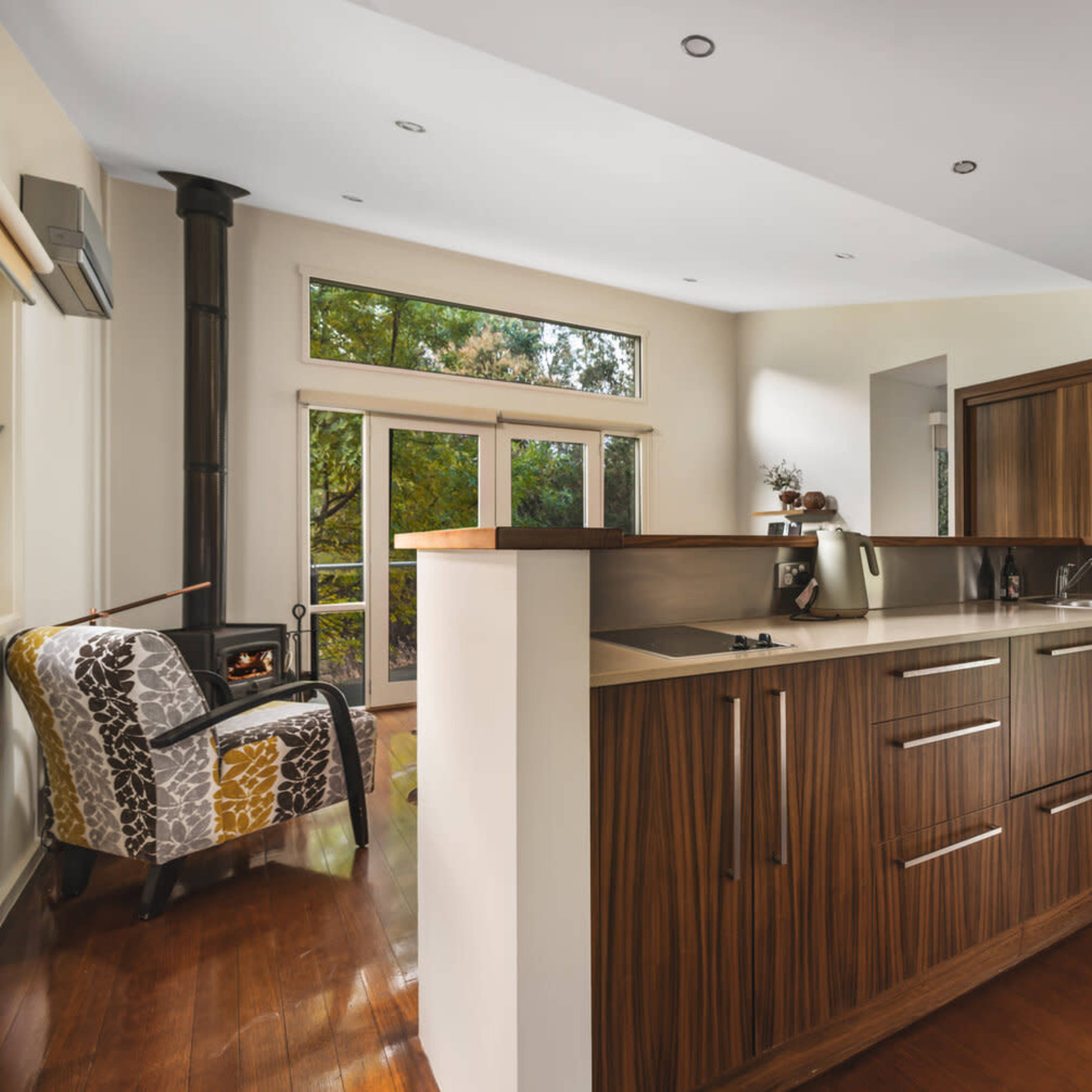 A modern kitchen with wooden cabinetry and a small dining area, featuring a stove and a view of outdoor greenery through large windows.