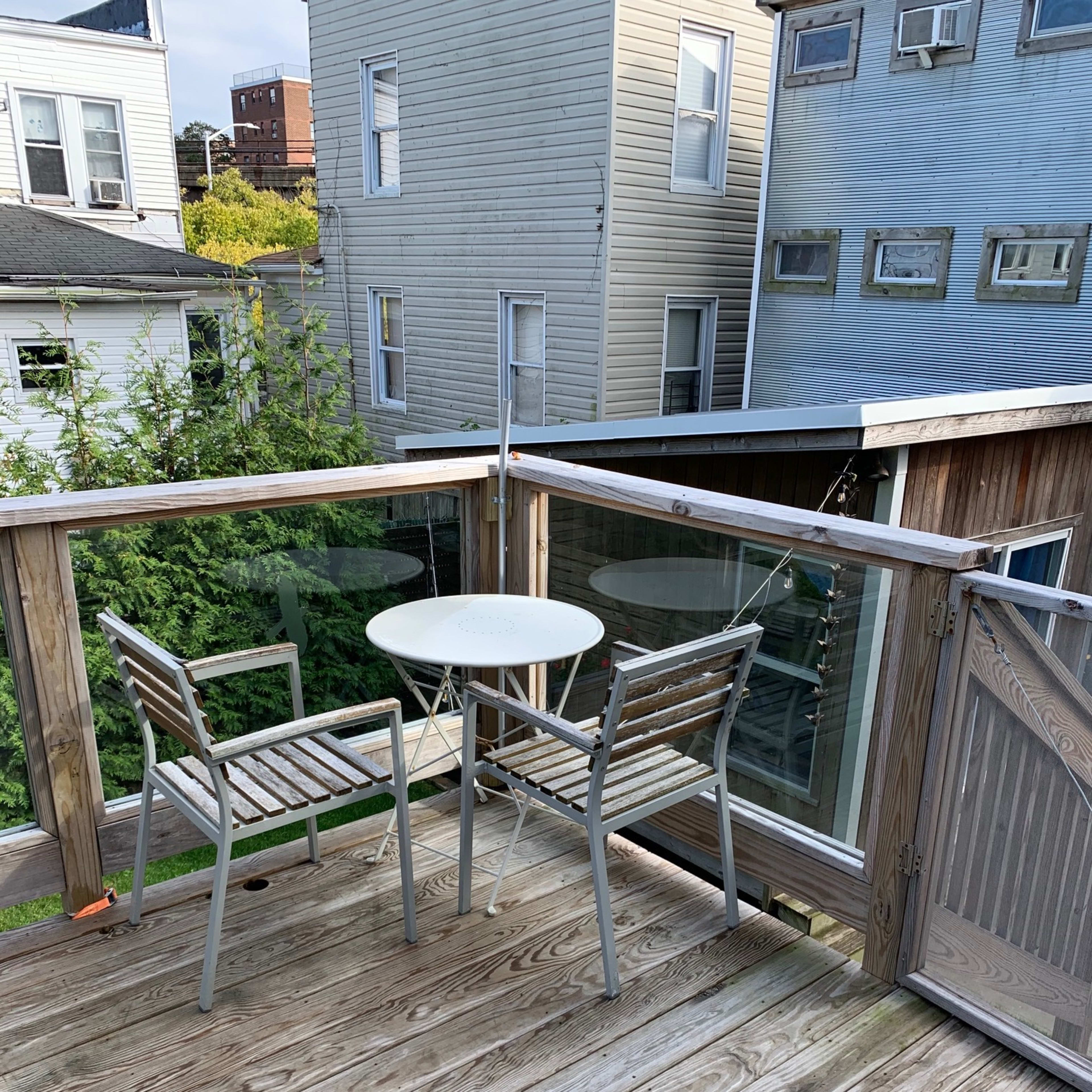 A small balcony with a round table and two wooden chairs, surrounded by wooden railings and facing neighboring buildings.