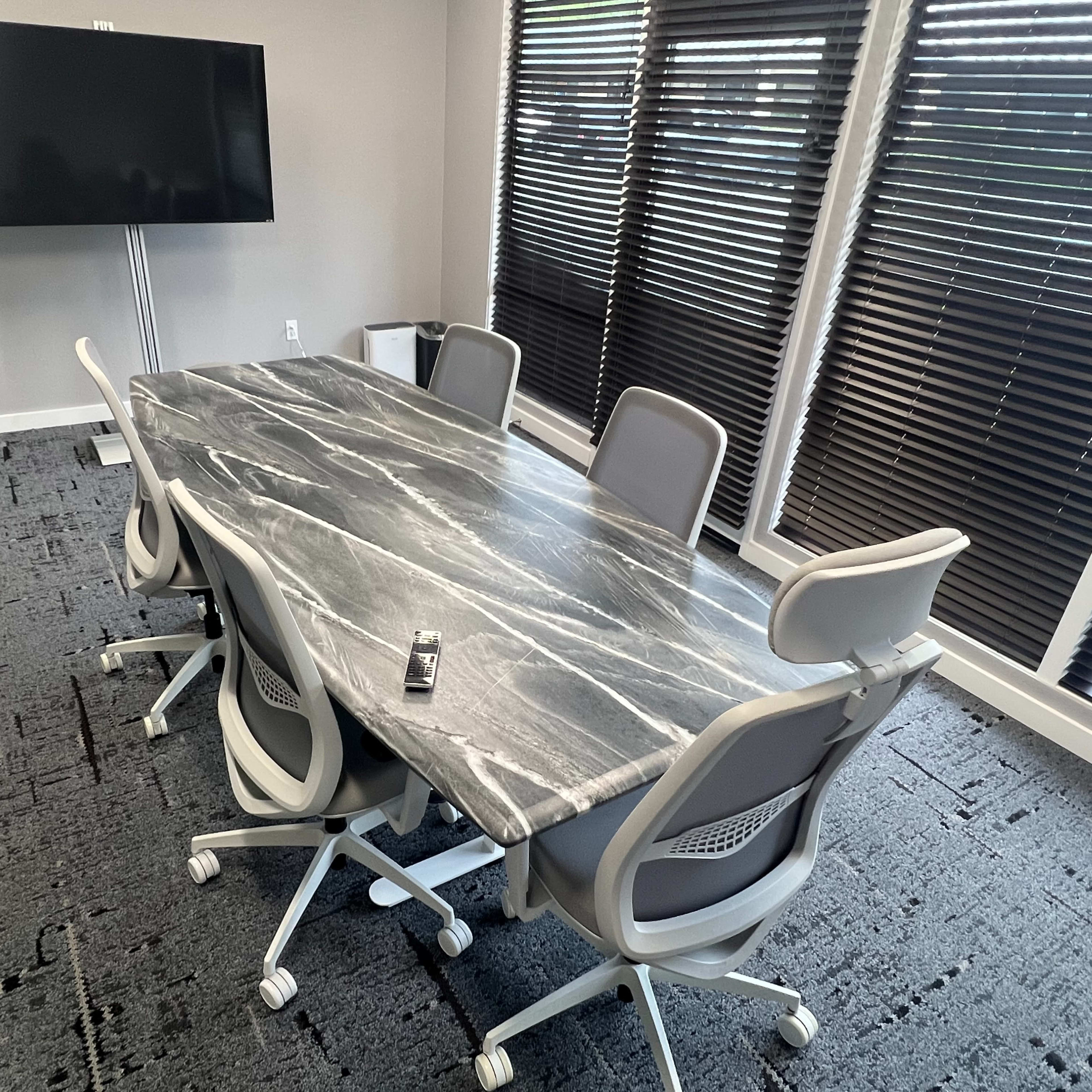 A marble conference table surrounded by five ergonomic chairs is situated in a brightly lit meeting room with large windows covered by blinds.