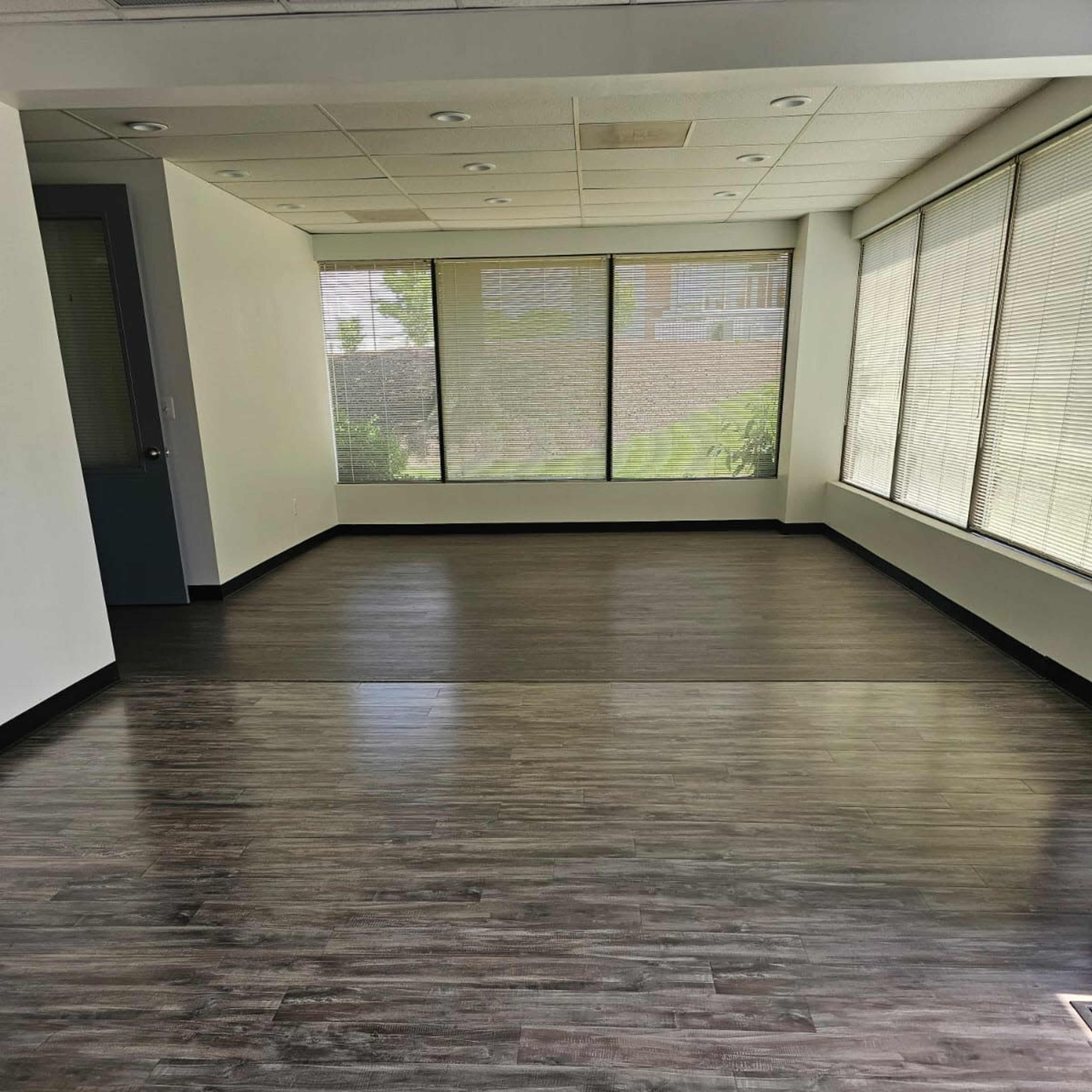An empty room with dark laminate flooring and large windows covered with blinds.
