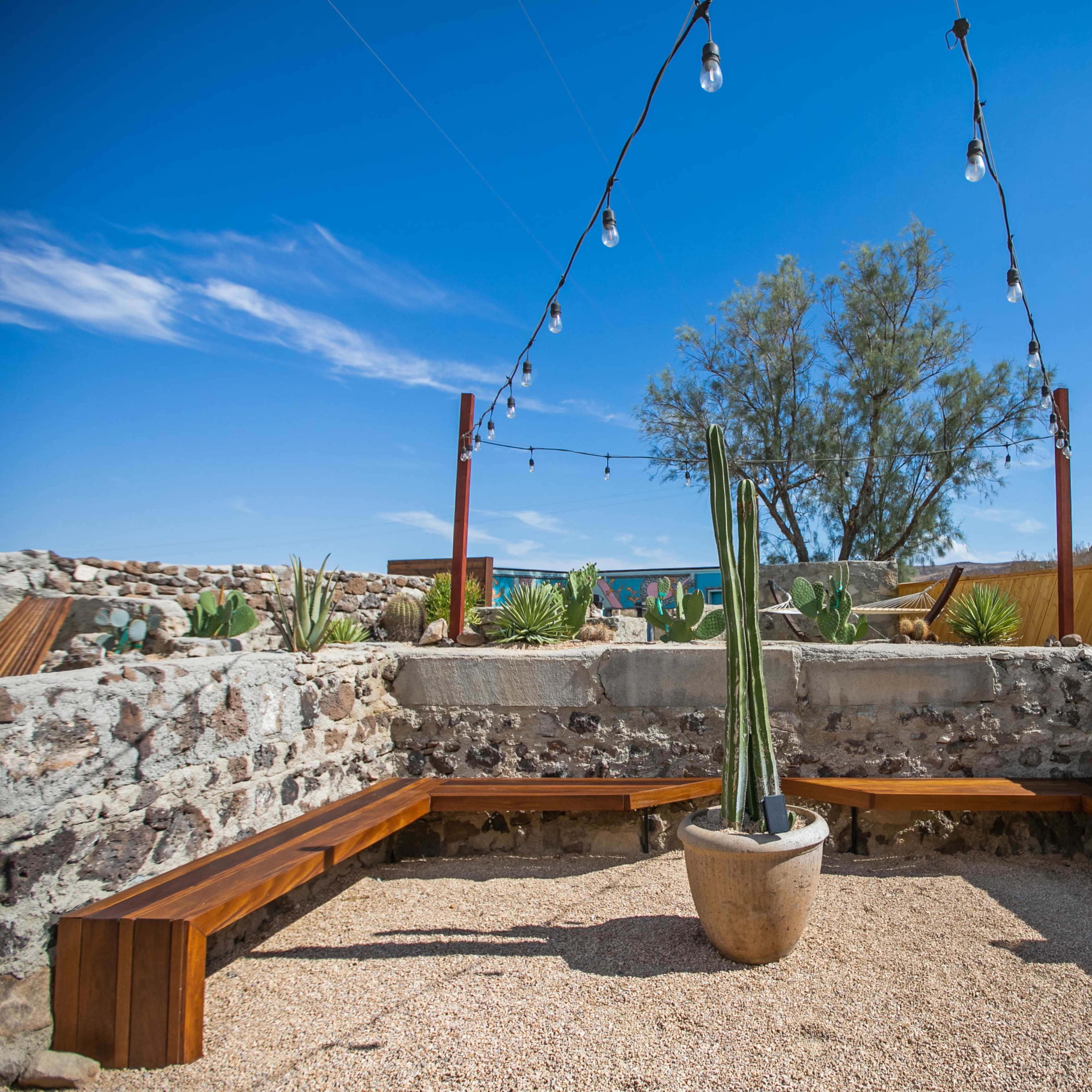 A circular seating area made of stone and wood surrounds a potted plant under string lights, set against a desert landscape.