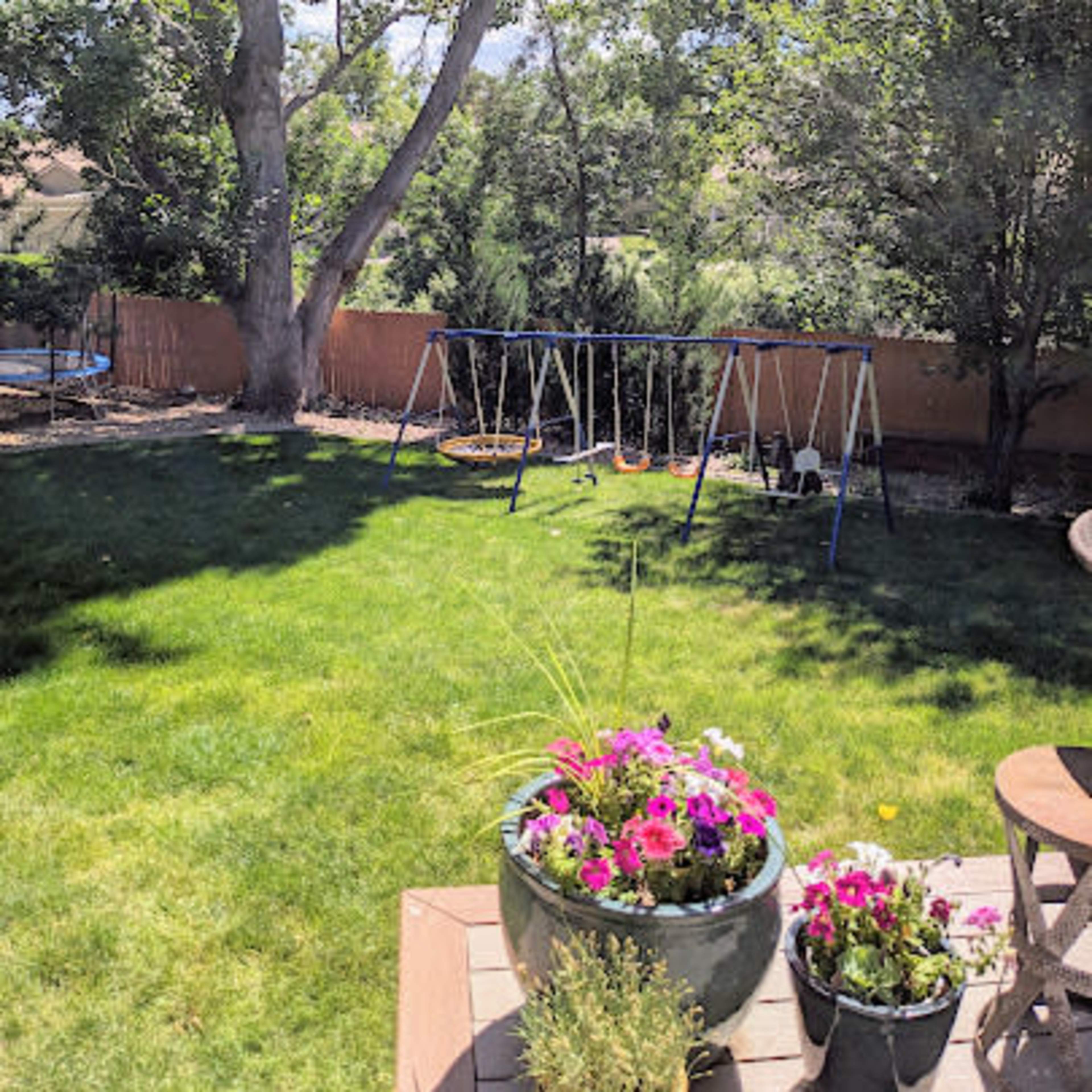 A grassy backyard features a swing set surrounded by trees and colorful flower pots in the foreground.