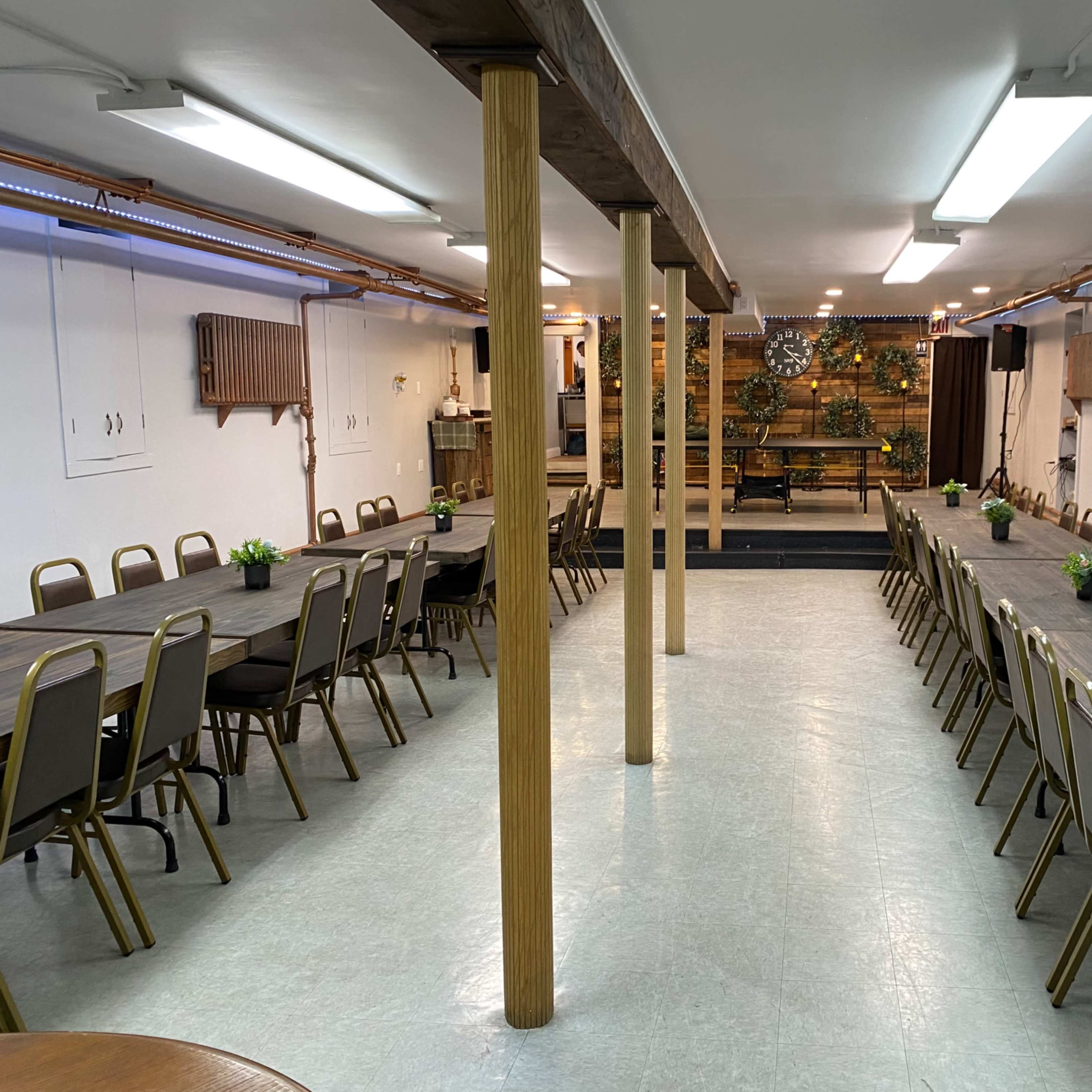 A large meeting room with long tables arranged in two parallel lines, surrounded by simple chairs and decorated with small plants on each table.