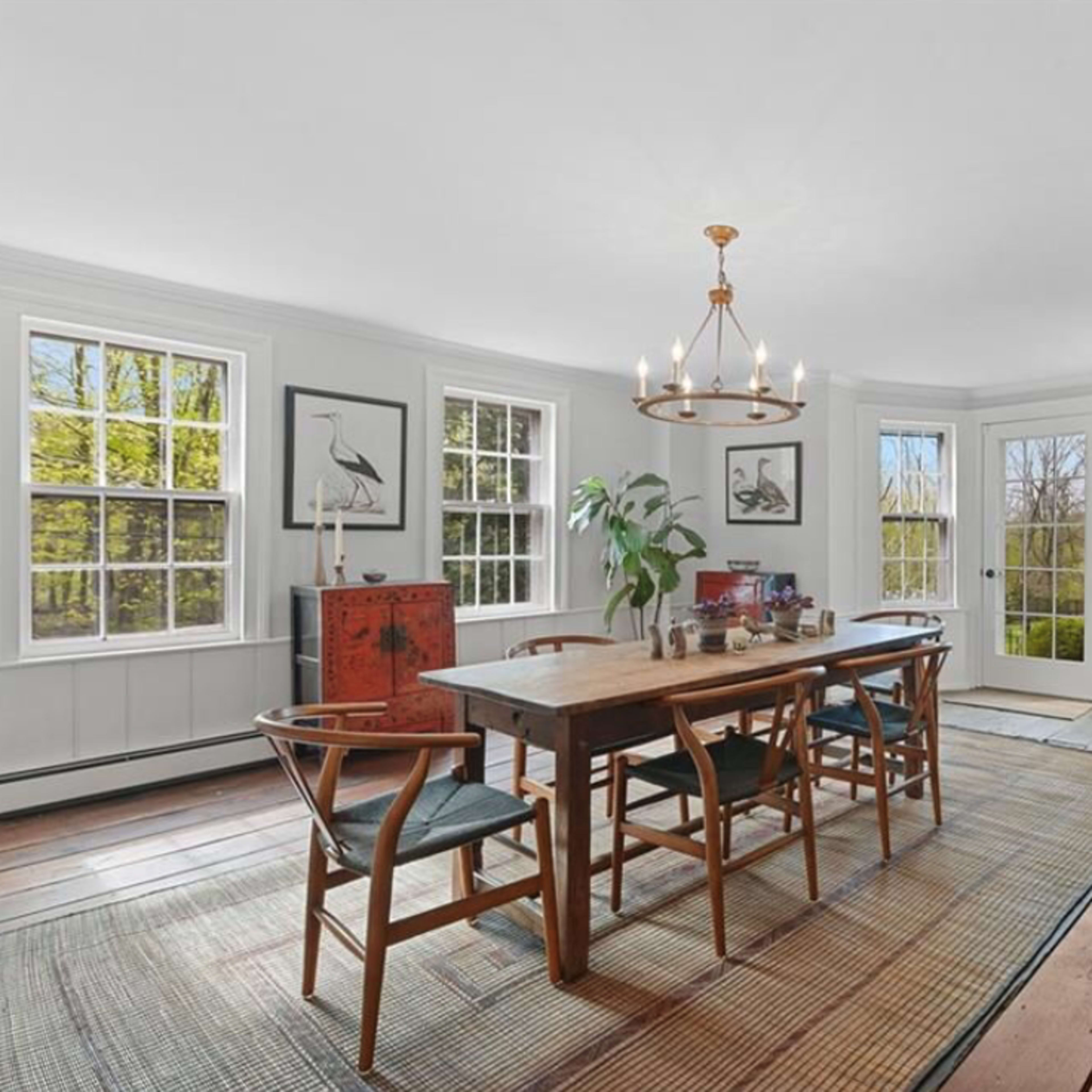 A dining room with a wooden table, six chairs, large windows, and a round mirror on the wall.