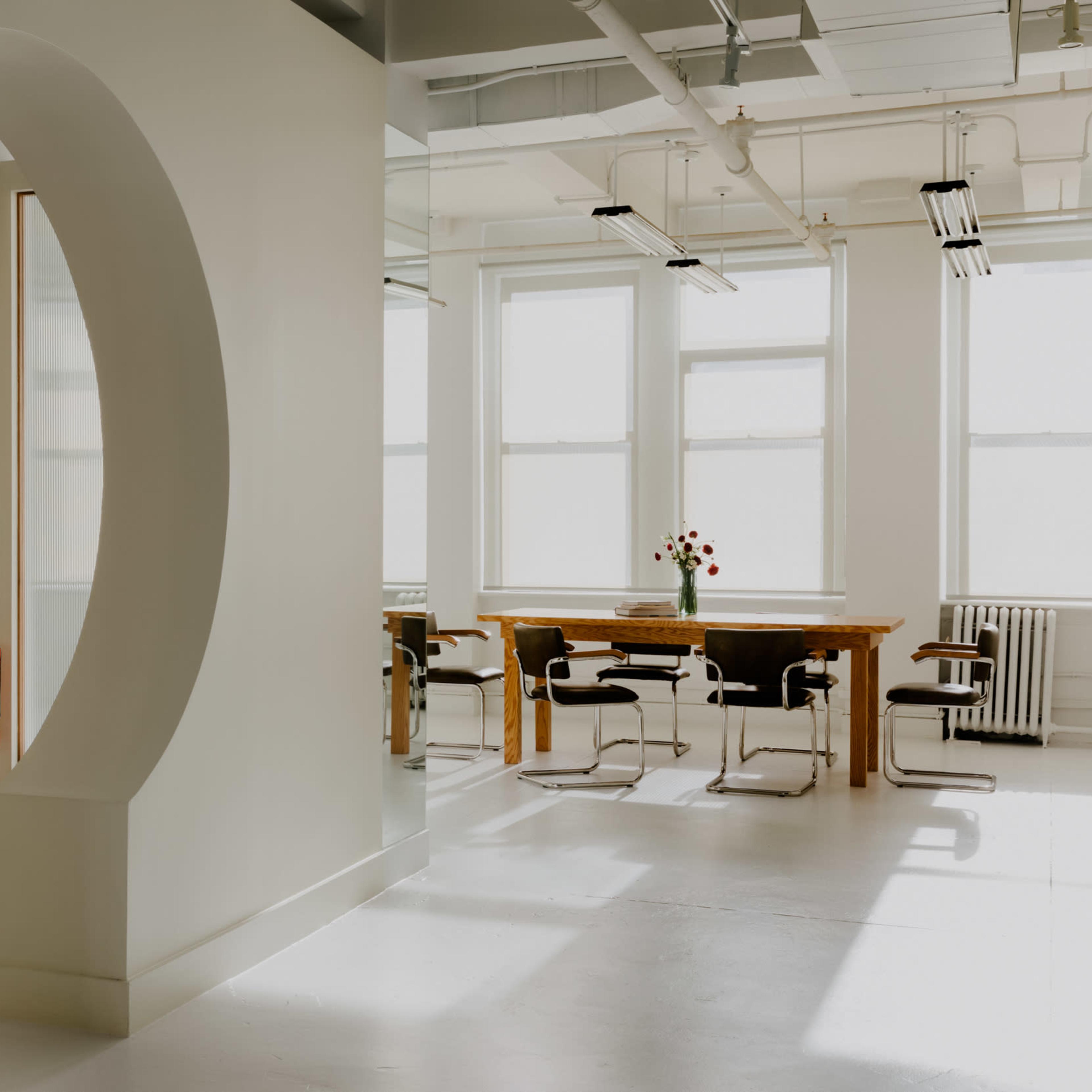 A modern office space features a circular opening leading to a cozy seating area, with a dining table and chairs visible in the background.