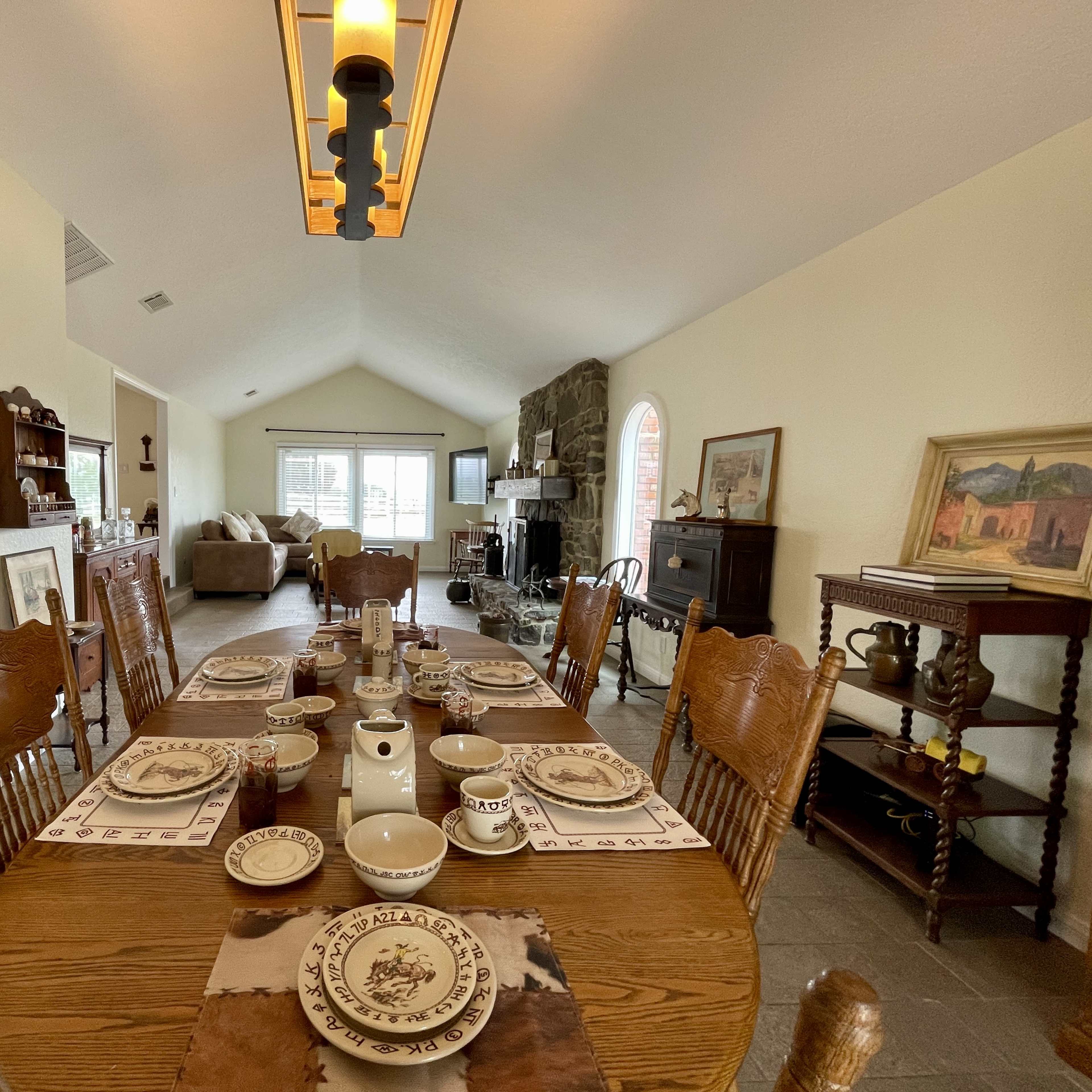 A dining table set for a meal with dishes and utensils is positioned in a spacious, open-concept living area featuring a stone fireplace and large windows.