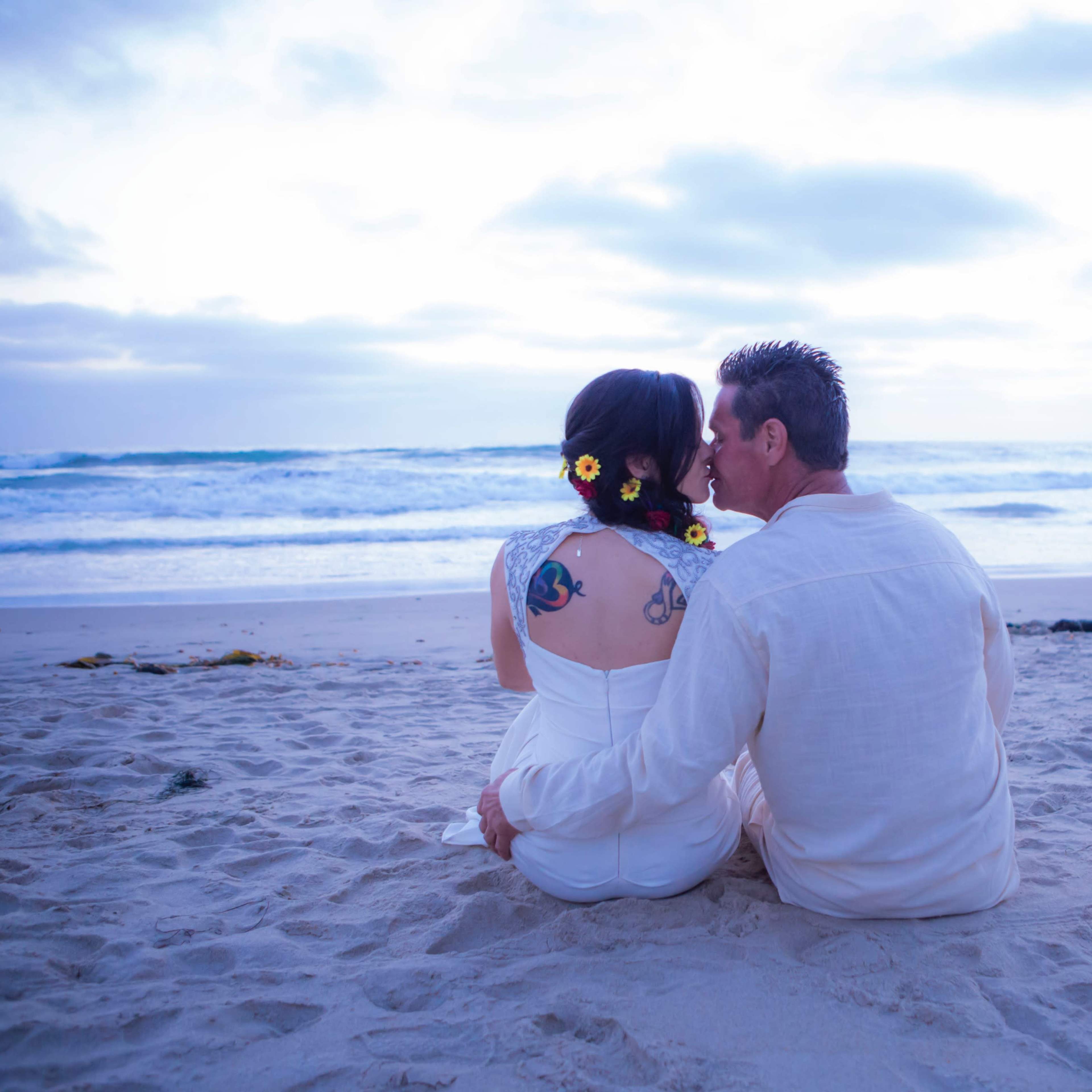 A couple sits on the beach, facing the ocean, sharing a moment as waves gently lap at the shore.