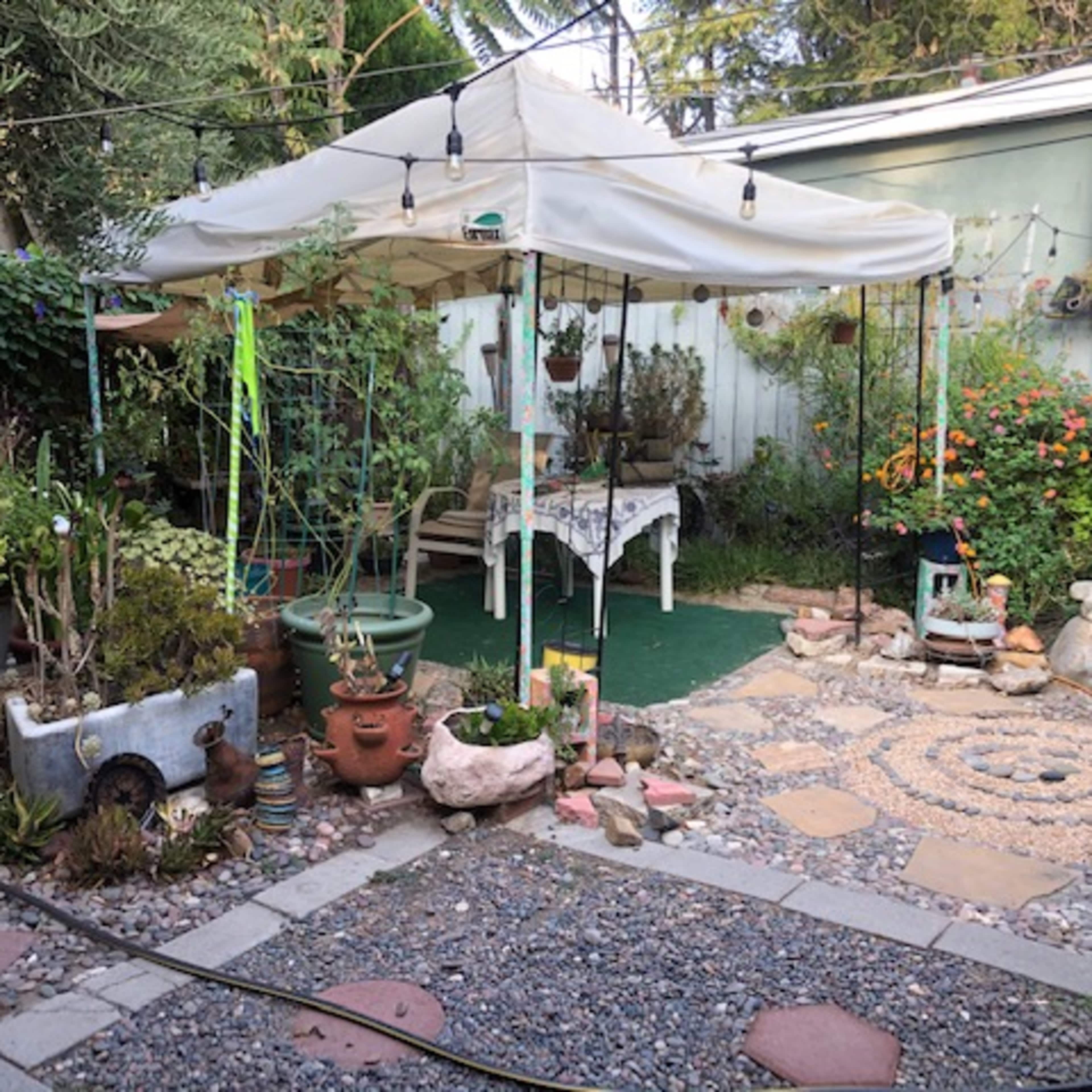 A garden space features a white canopy, various potted plants, a stone pathway, and decorative elements arranged around a circular stone pattern.