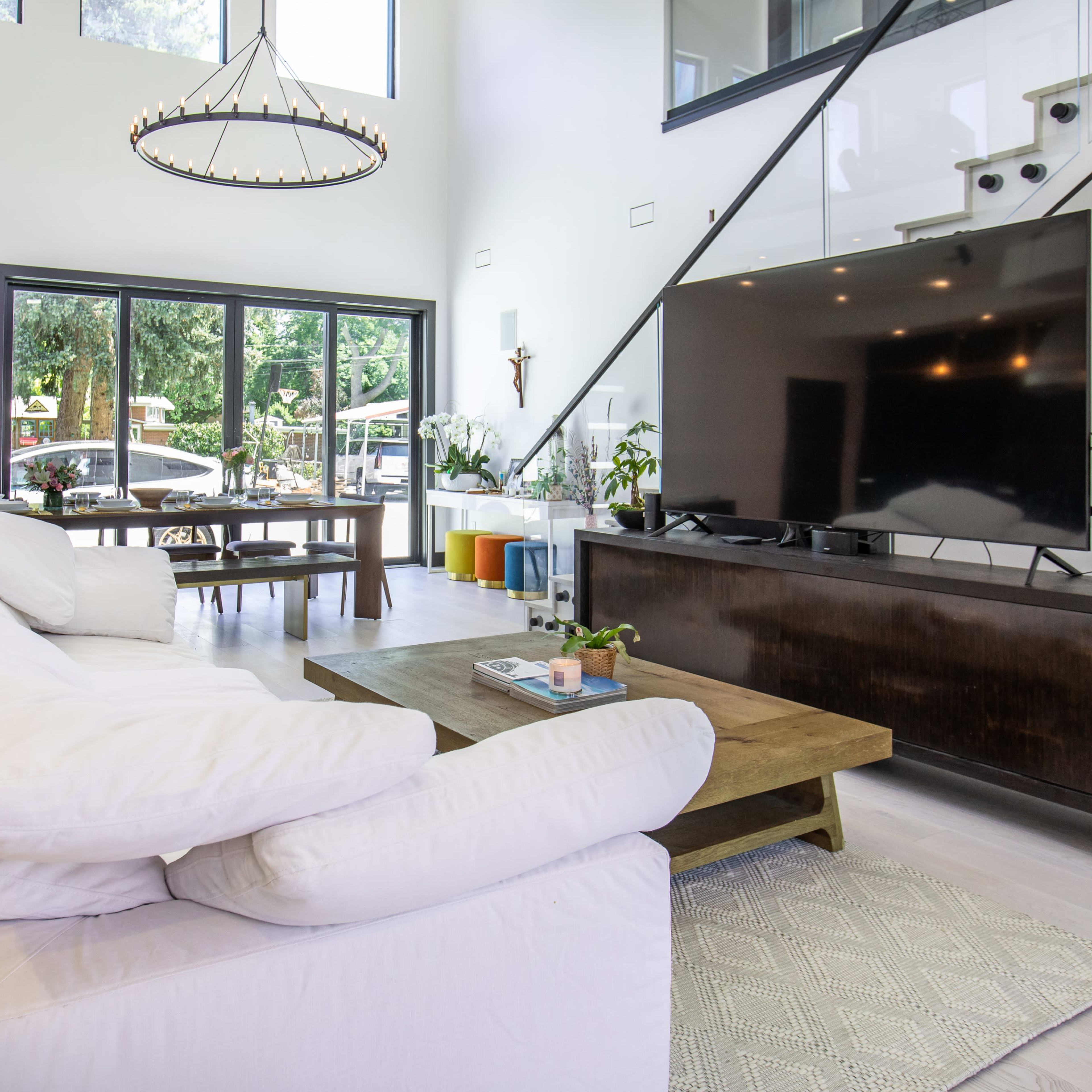 A modern living room featuring a white sectional sofa, a large flat-screen TV on a dark wooden console, and large windows that provide a view of the outdoors.