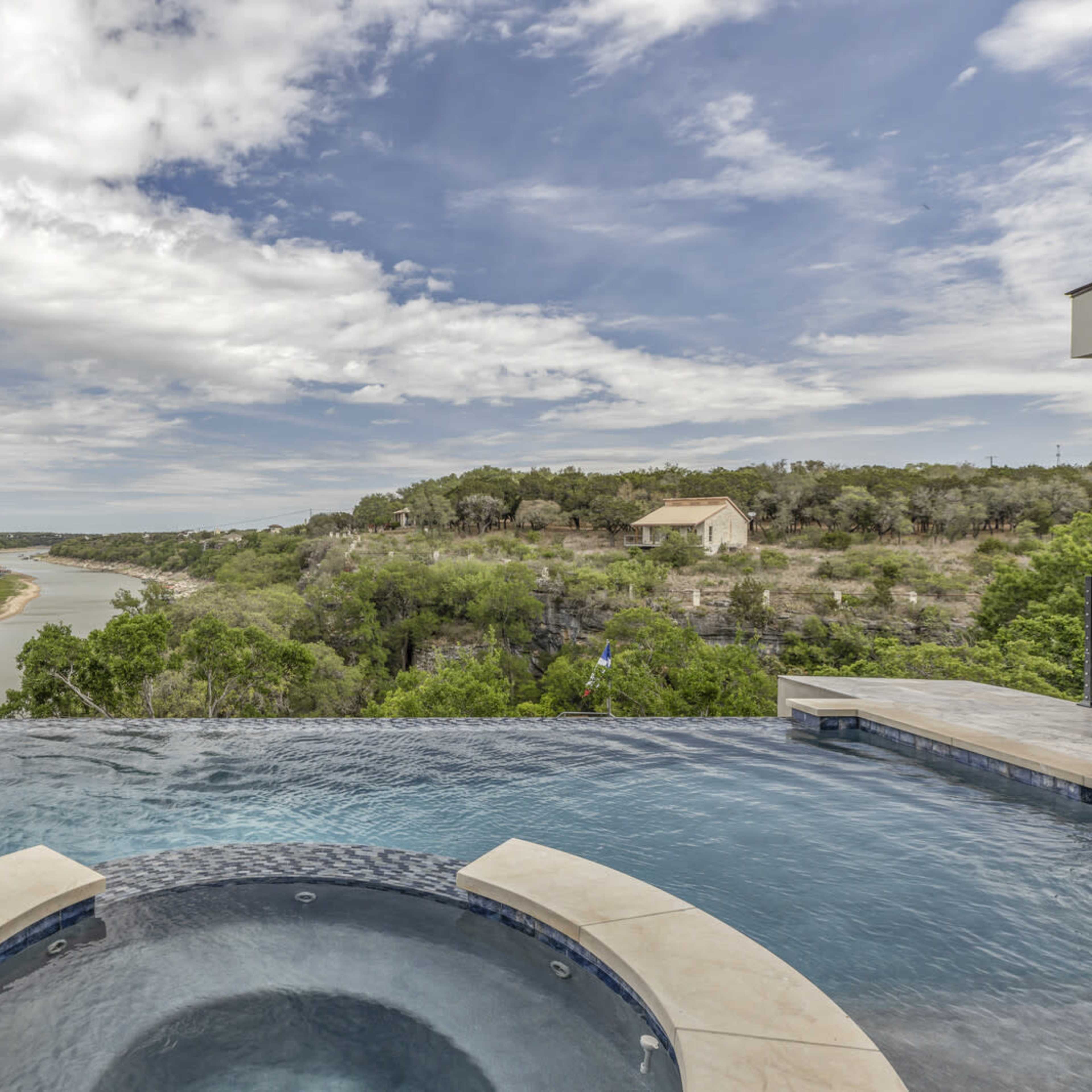 An infinity pool overlooks a winding river surrounded by lush greenery under a cloudy sky.