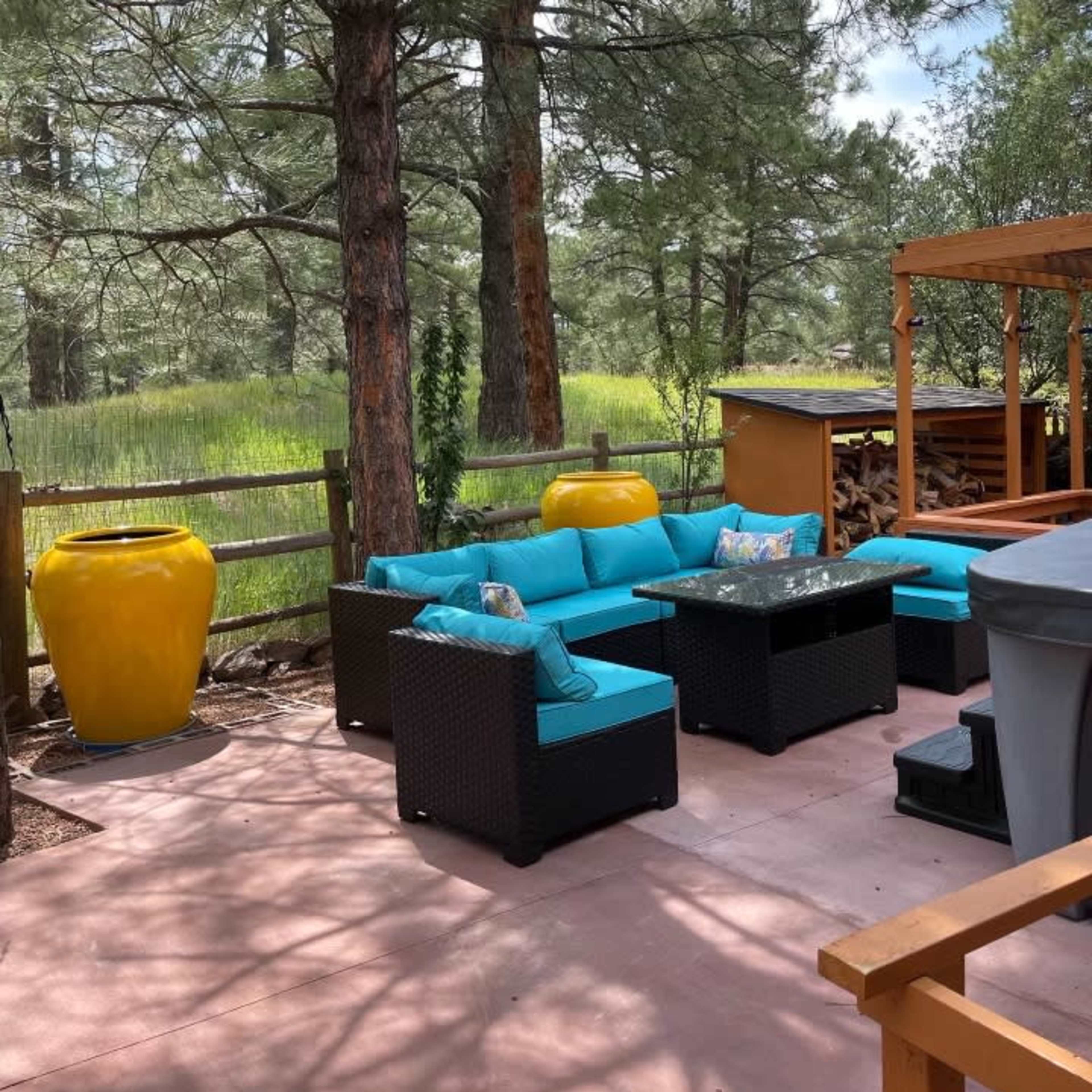 A patio area features turquoise furniture surrounded by tall pine trees and a wooden fence, with a yellow ceramic pot and a storage unit in the background.