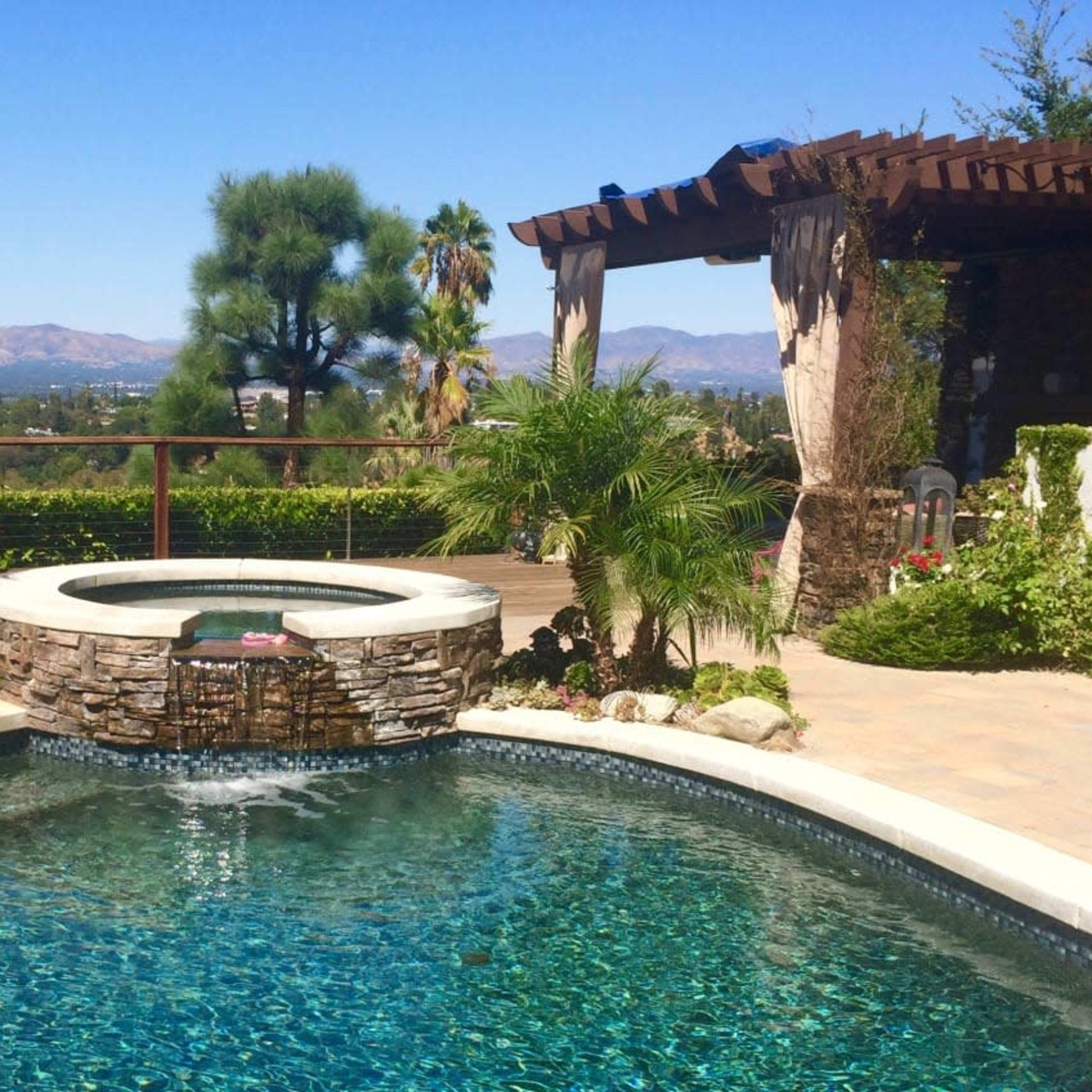 A swimming pool with a built-in spa and a stone waterfall is surrounded by tropical plants and offers a view of distant mountains under a clear blue sky.