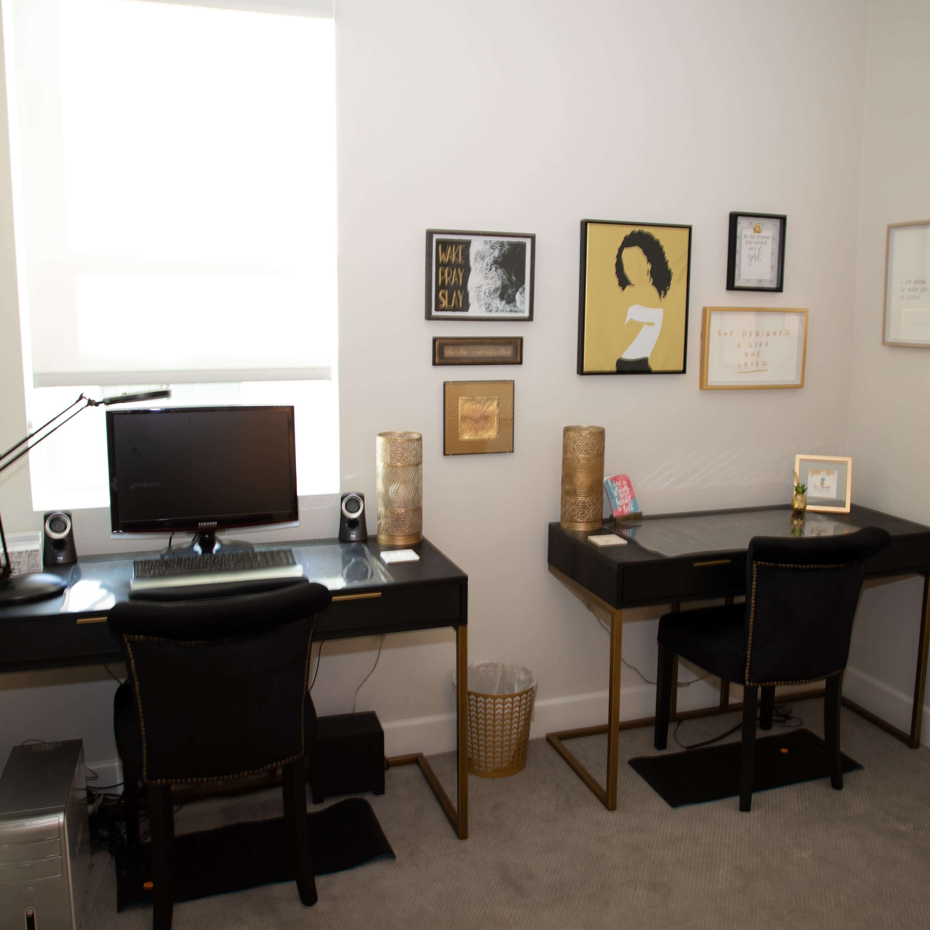 A home office with two black desks, each equipped with a chair and decorative wall art, illuminated by natural light from a window.