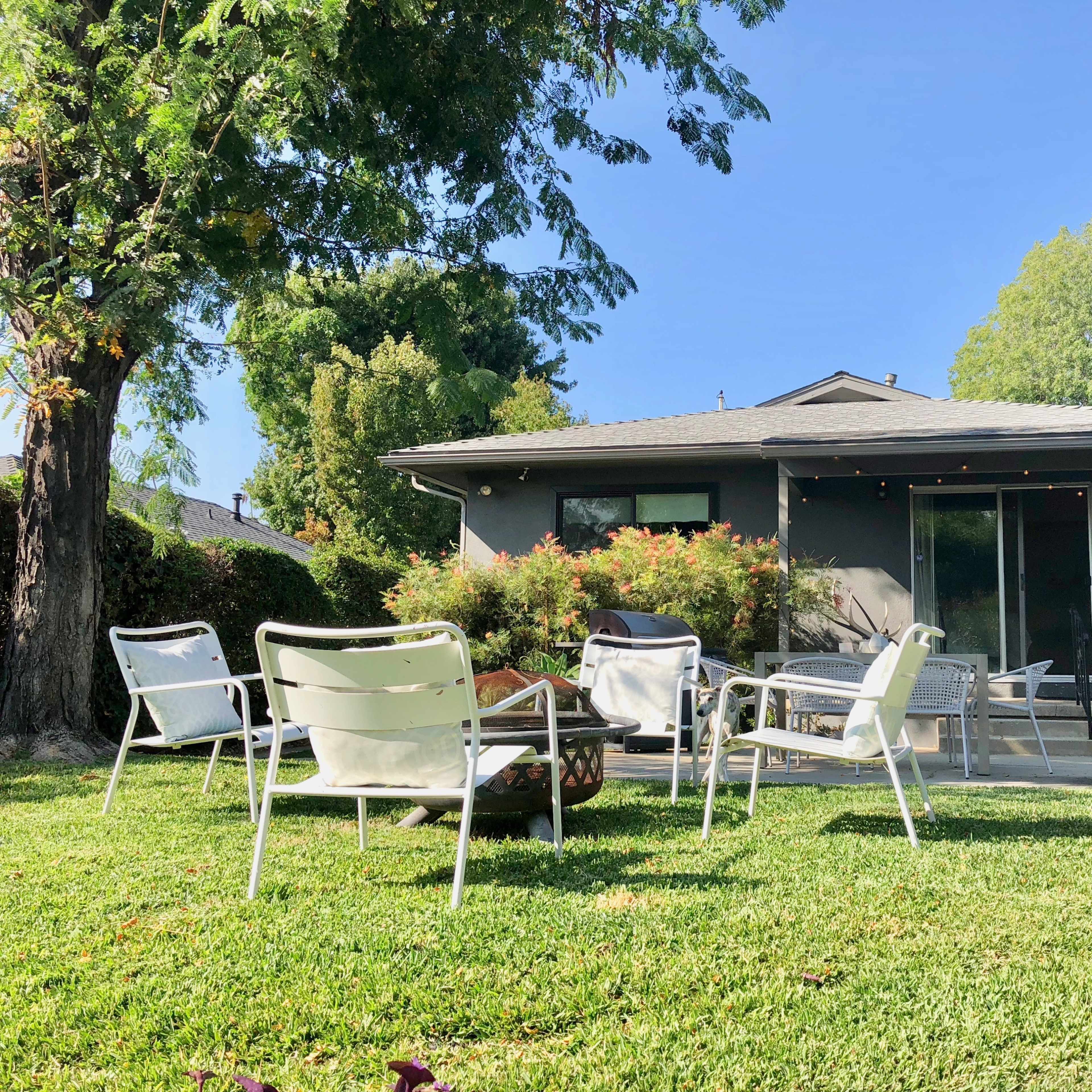 A modern house with a backyard surrounded by chairs and greenery, featuring sliding glass doors and string lights overhead.