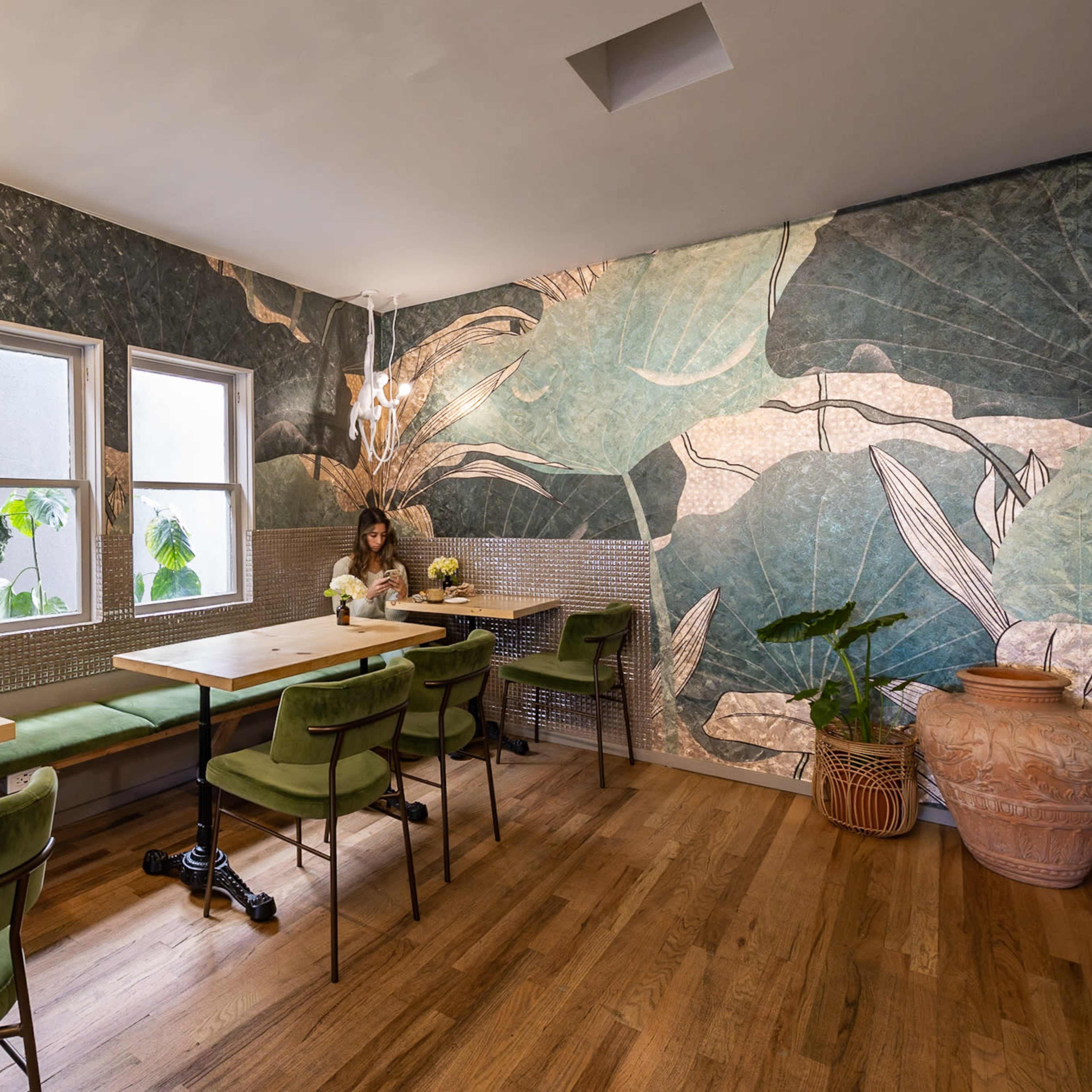 A woman sits alone at a wooden table in a cafe with leaf-patterned wallpaper, two windows, and green velvet chairs.