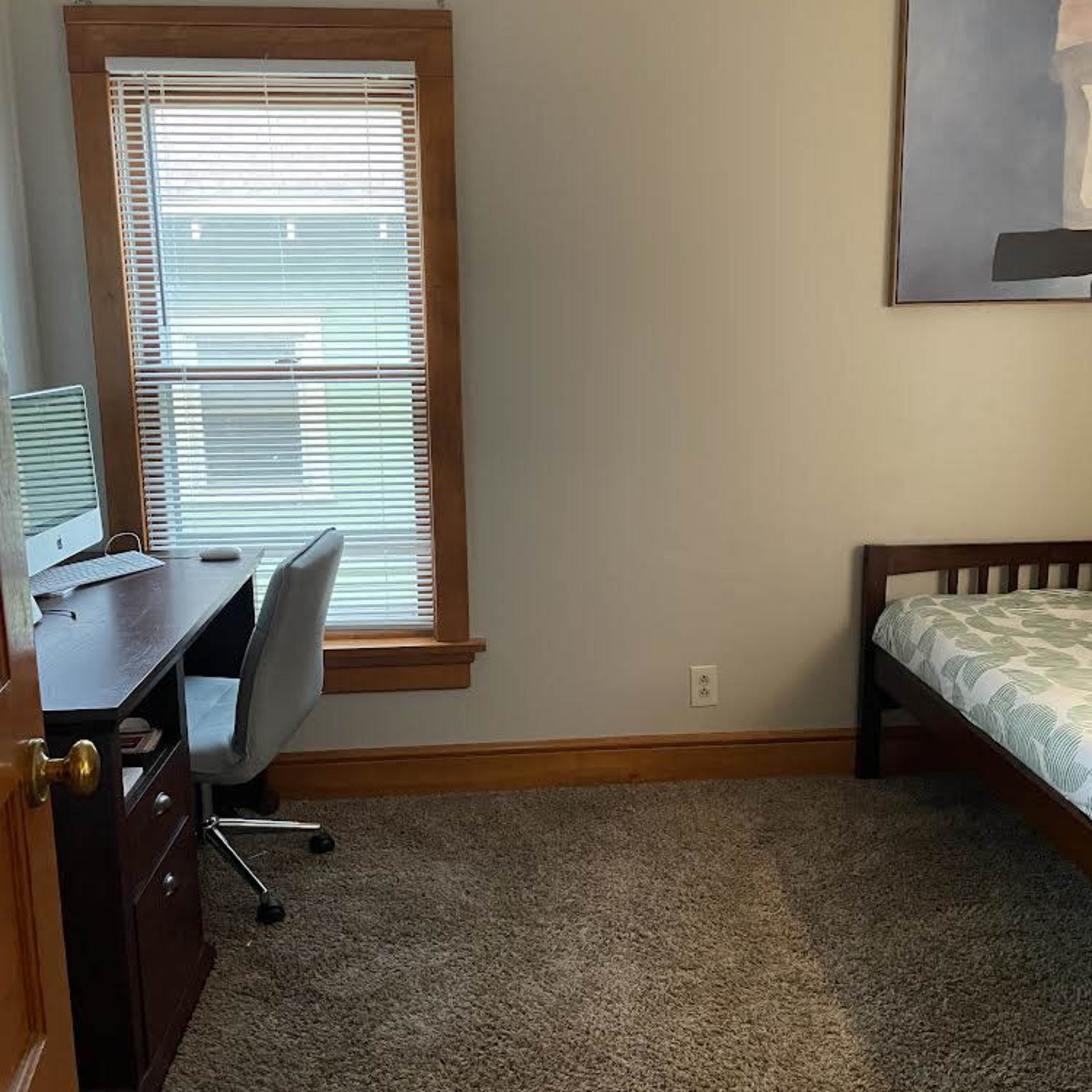 A small room with a bed, a desk with a computer, and a window with blinds.