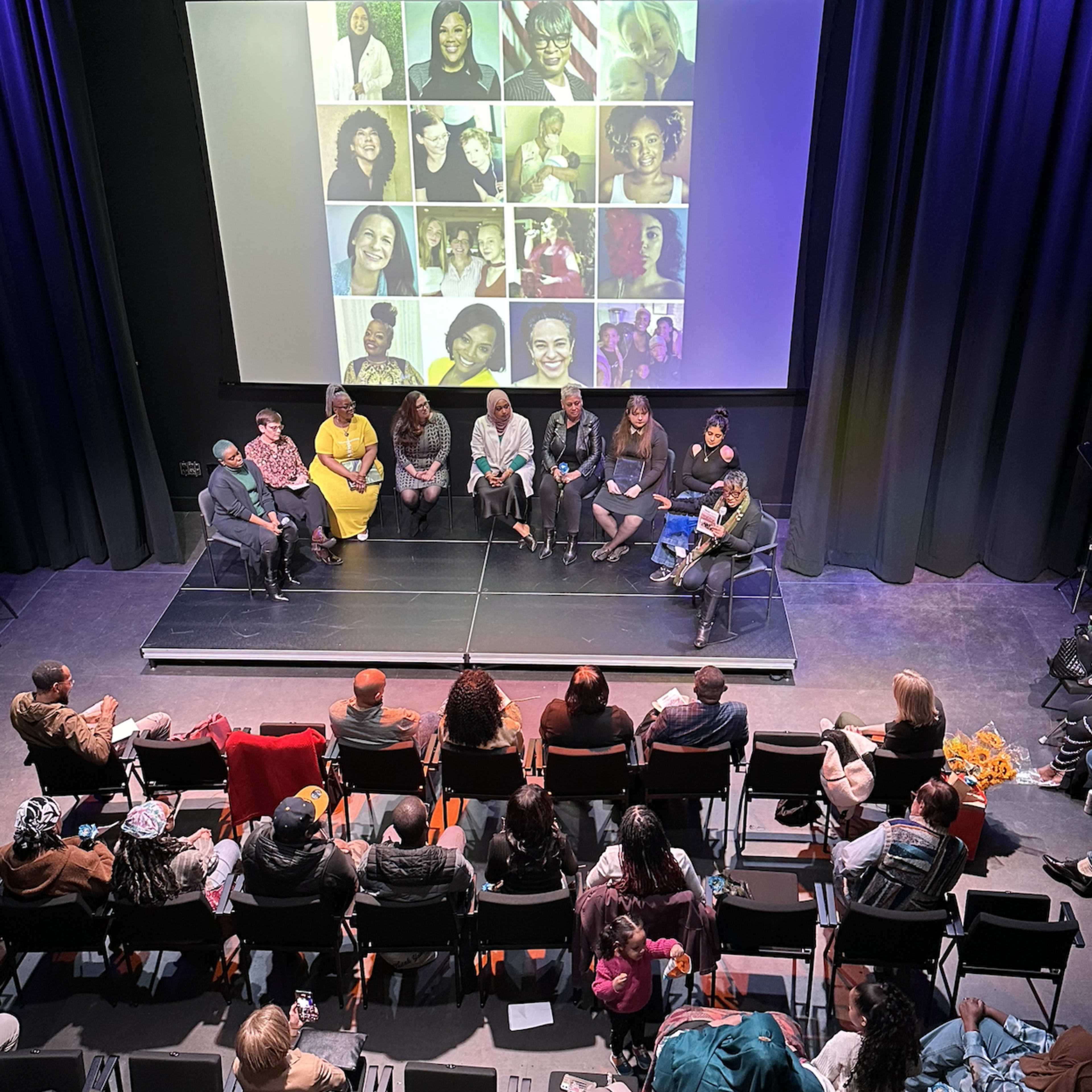 A panel of speakers sits on a stage with a large screen displaying photos behind them, while an audience watches from seated rows.