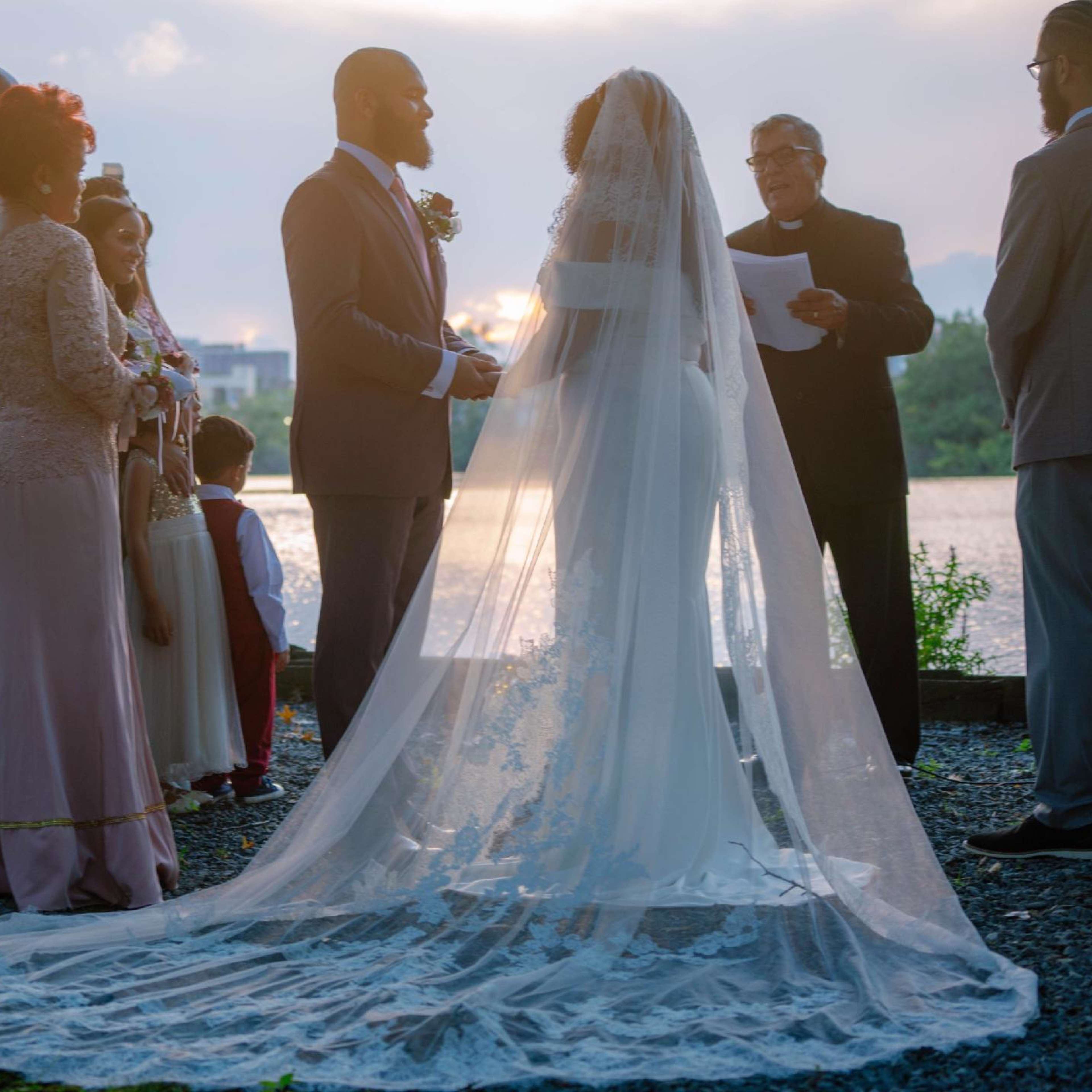 A couple stands under a tree while exchanging vows during a wedding ceremony by the water, surrounded by family and friends.