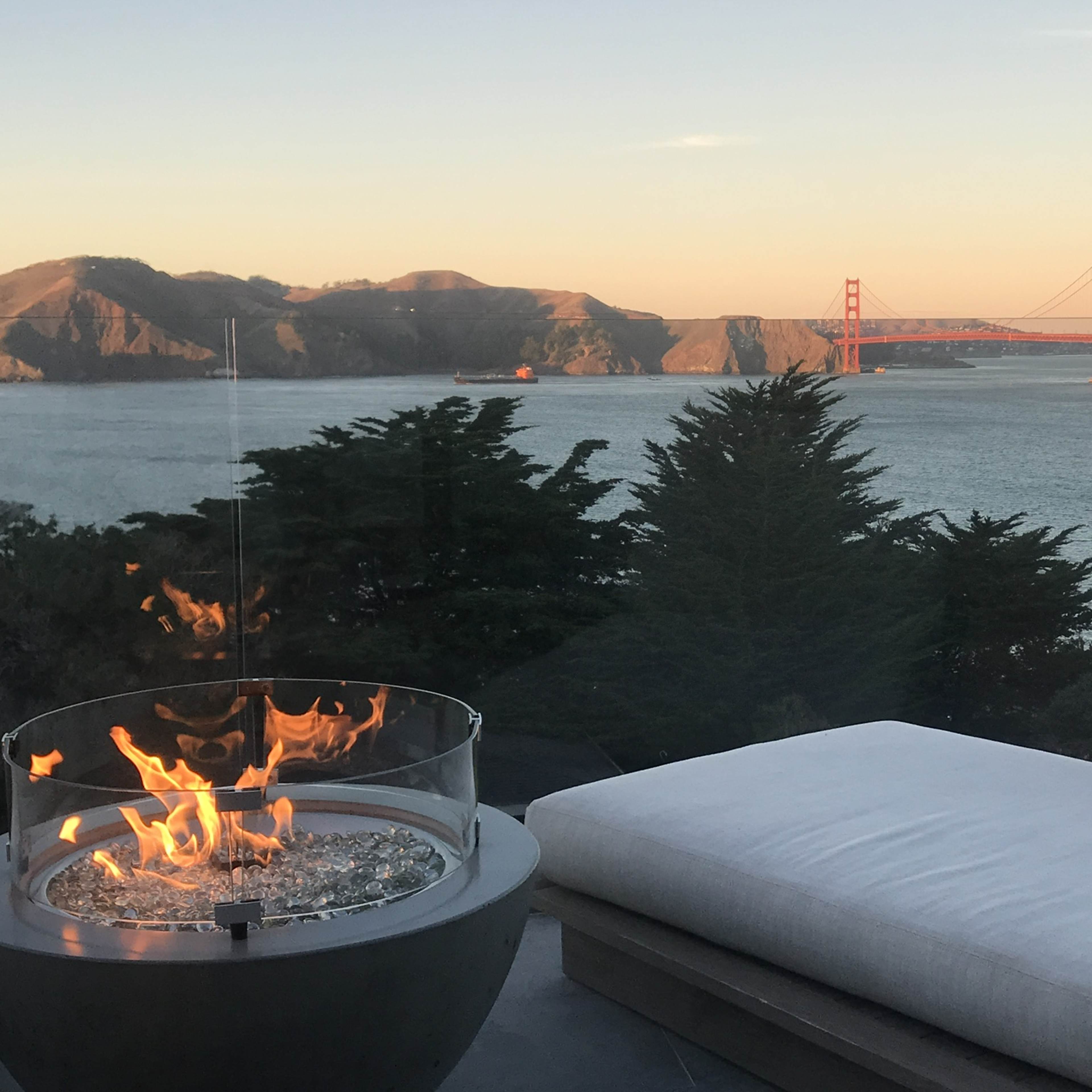 A modern seating area features a circular fire pit with flames, overlooking the Golden Gate Bridge and surrounding hills at sunset.