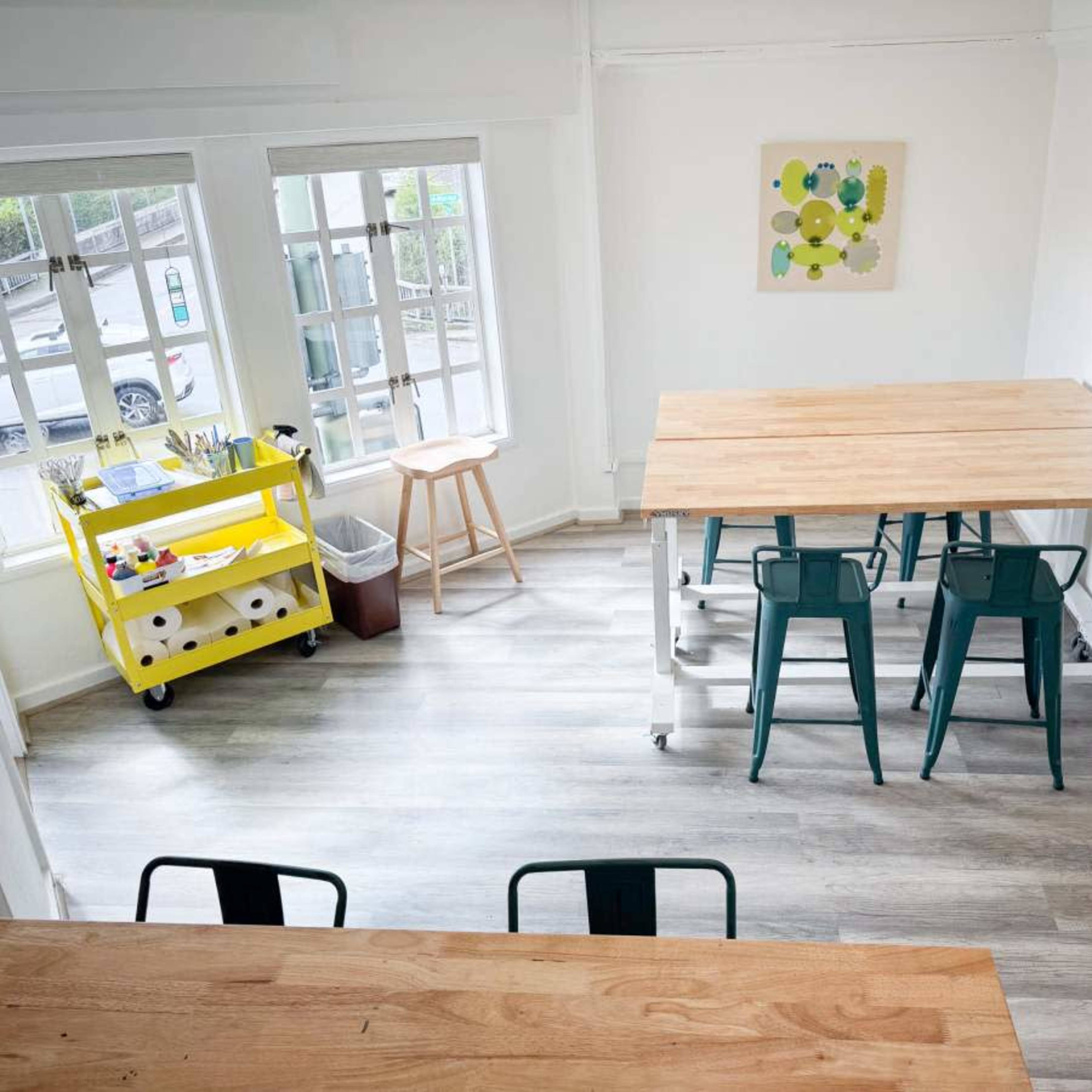 A bright room with wooden tables, green metal chairs, and large windows overlooking parked cars.