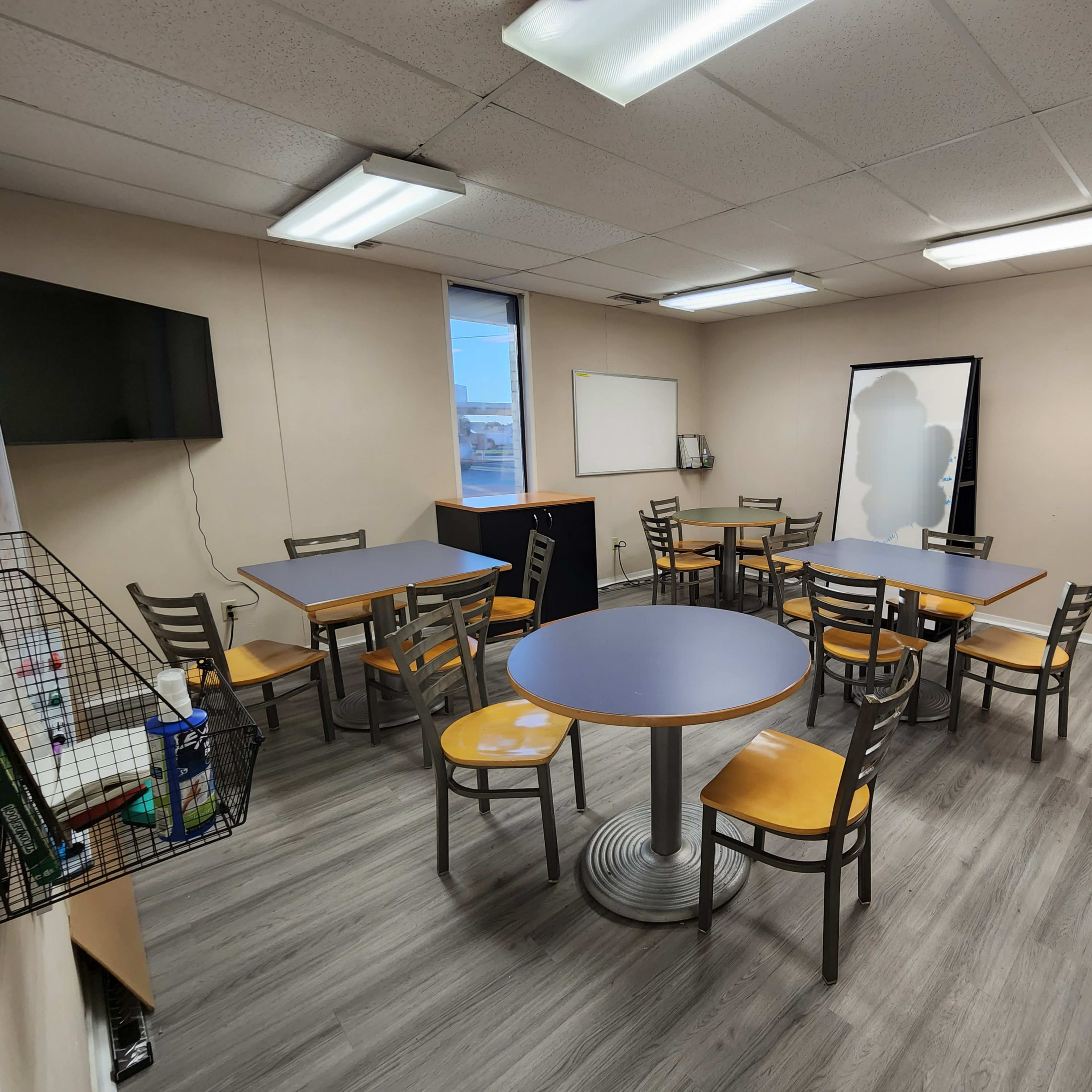 A well-lit room with multiple tables and chairs arranged for collaborative seating, featuring a TV, a whiteboard, and storage cabinets.