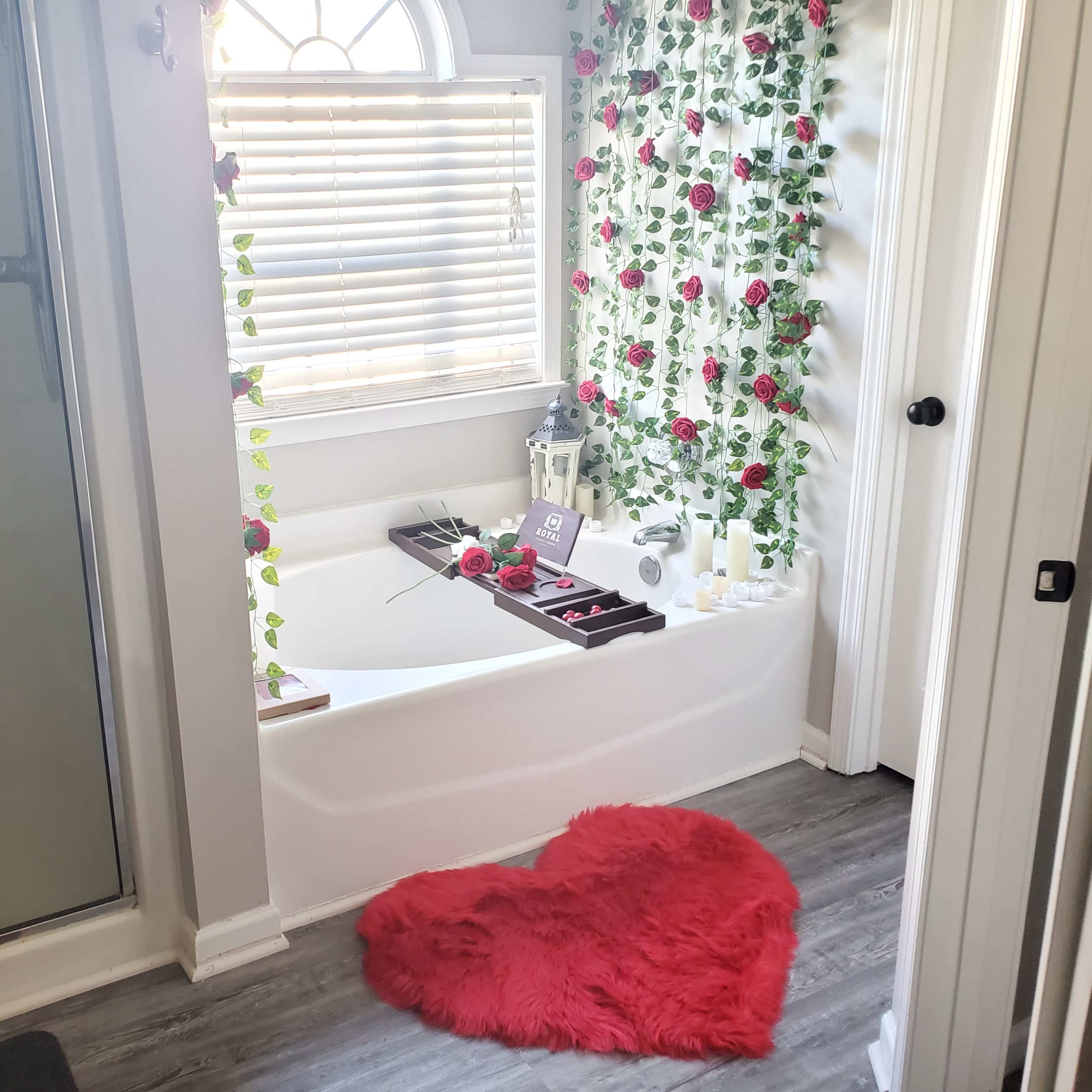 A bathroom features a white bathtub adorned with rose decorations, a heart-shaped rug, and a nearby shower area with glass doors.