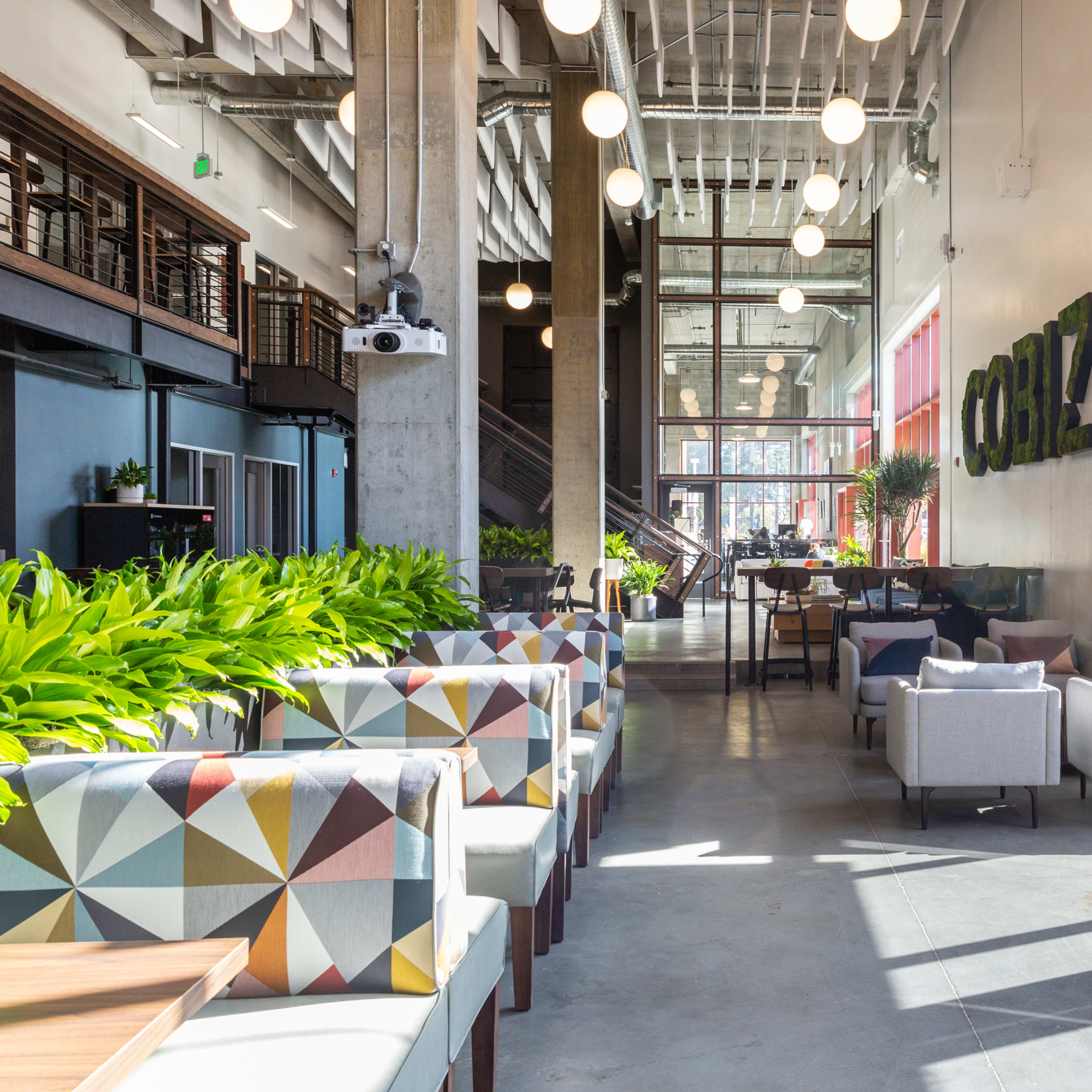 A modern café interior with colorful seating, large windows, and greenery throughout.