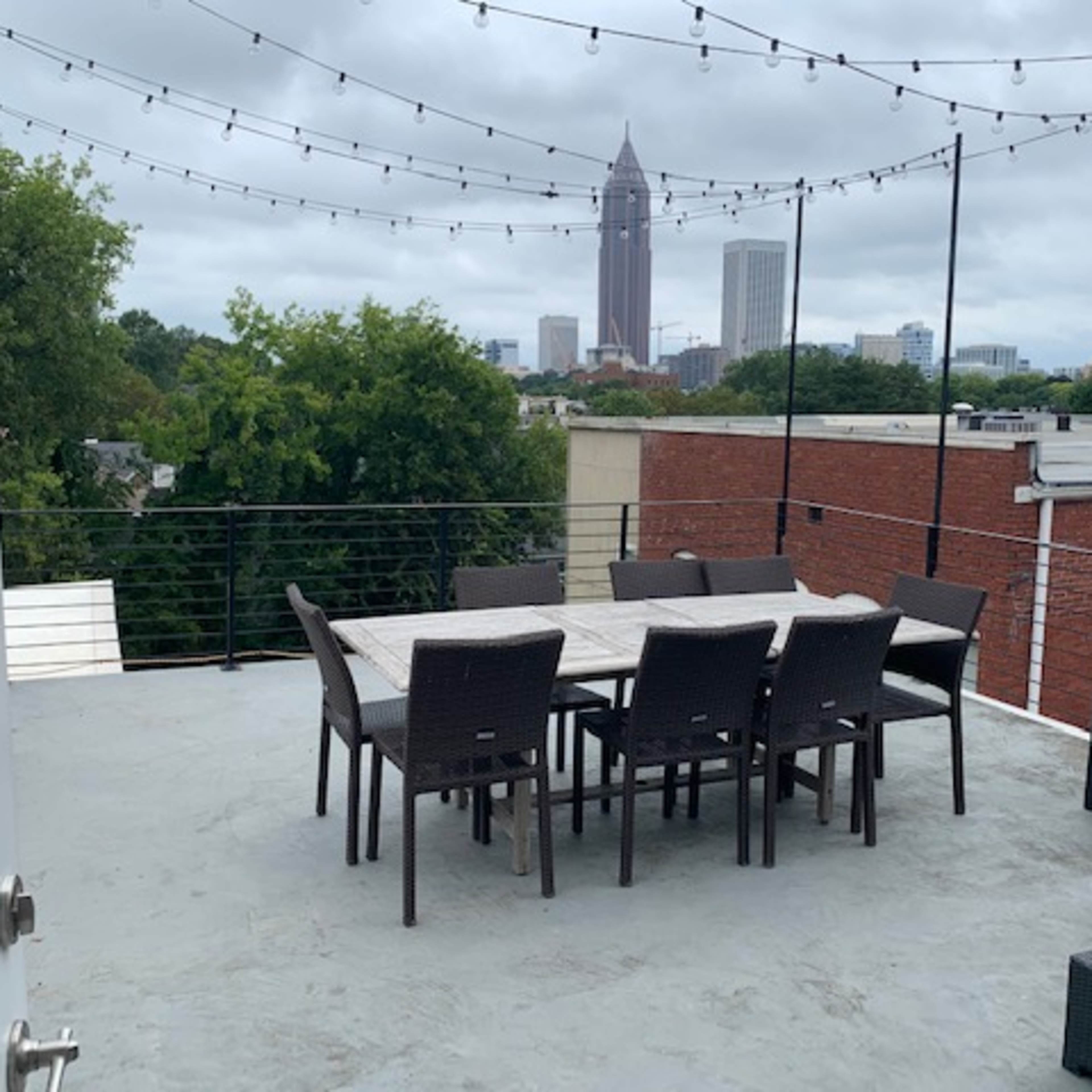 A rooftop patio features a dining table surrounded by chairs and a view of tall buildings in the background under a cloudy sky.