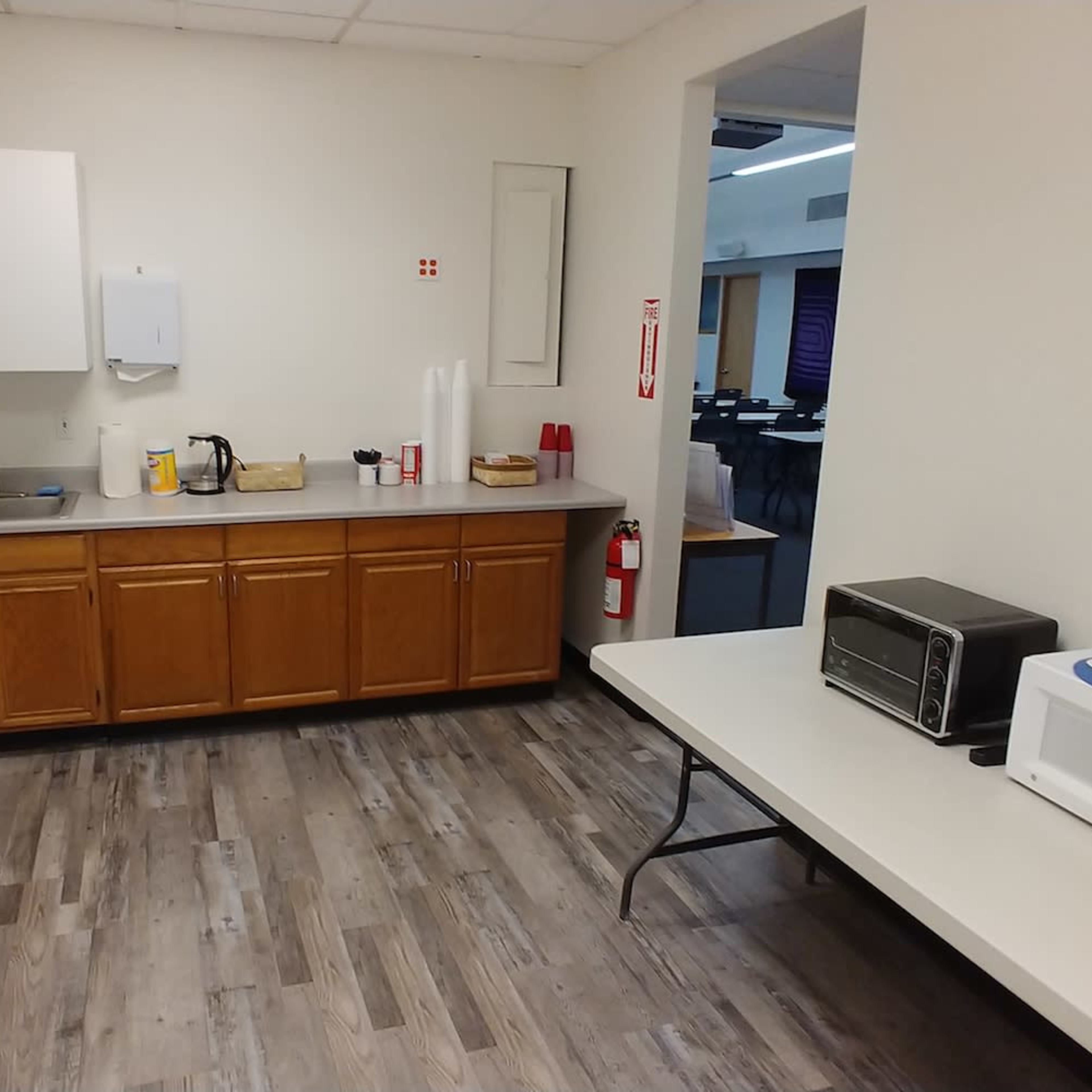 A break room with a kitchenette featuring cabinets, a sink, and appliances, including a microwave and a coffee maker, alongside a foldable table and a trash bin.