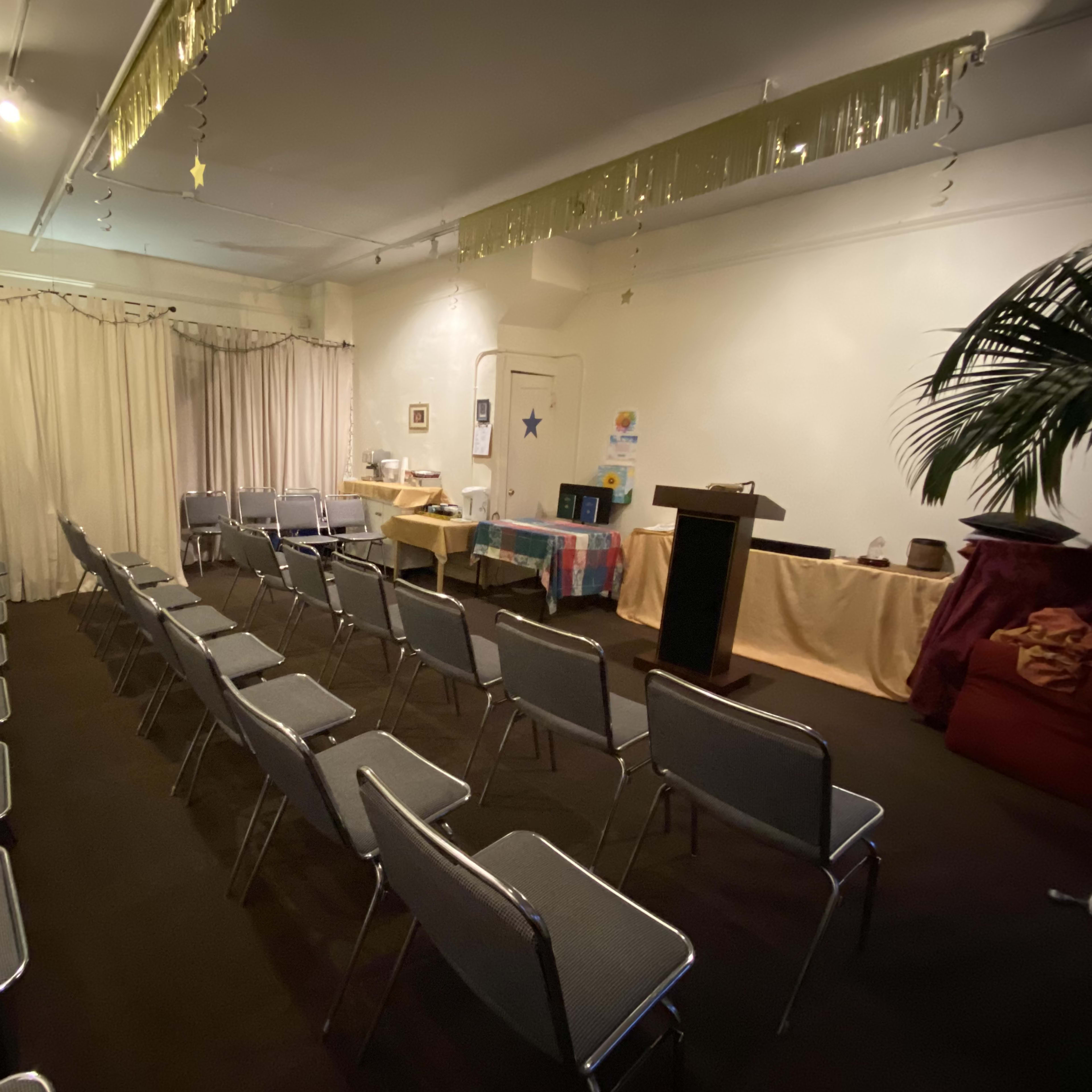 A room with neatly arranged metal chairs facing a small podium, surrounded by tables and decorations, including a large curtain and a potted plant.