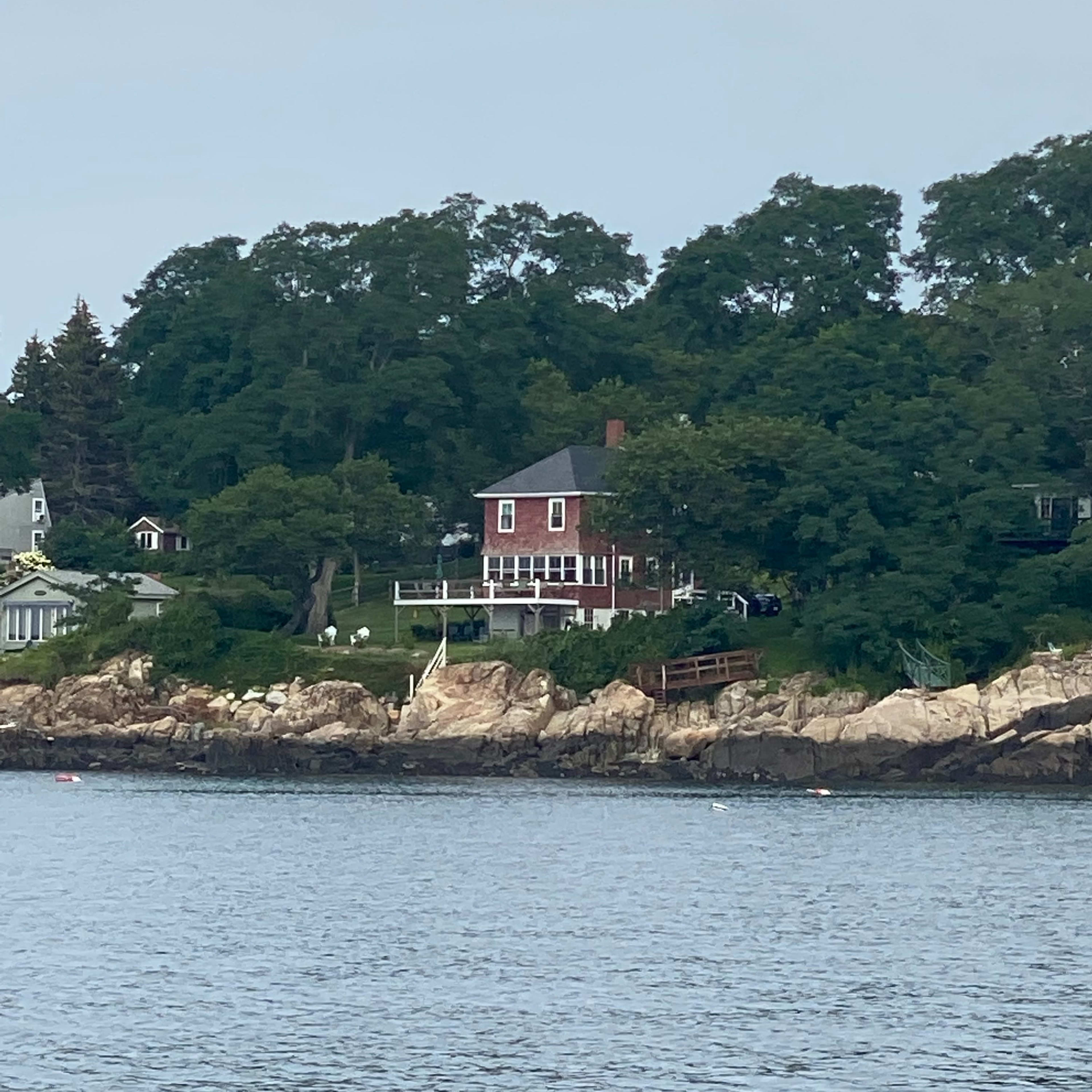A red house is situated on a rocky shoreline, surrounded by trees and overlooking the water.