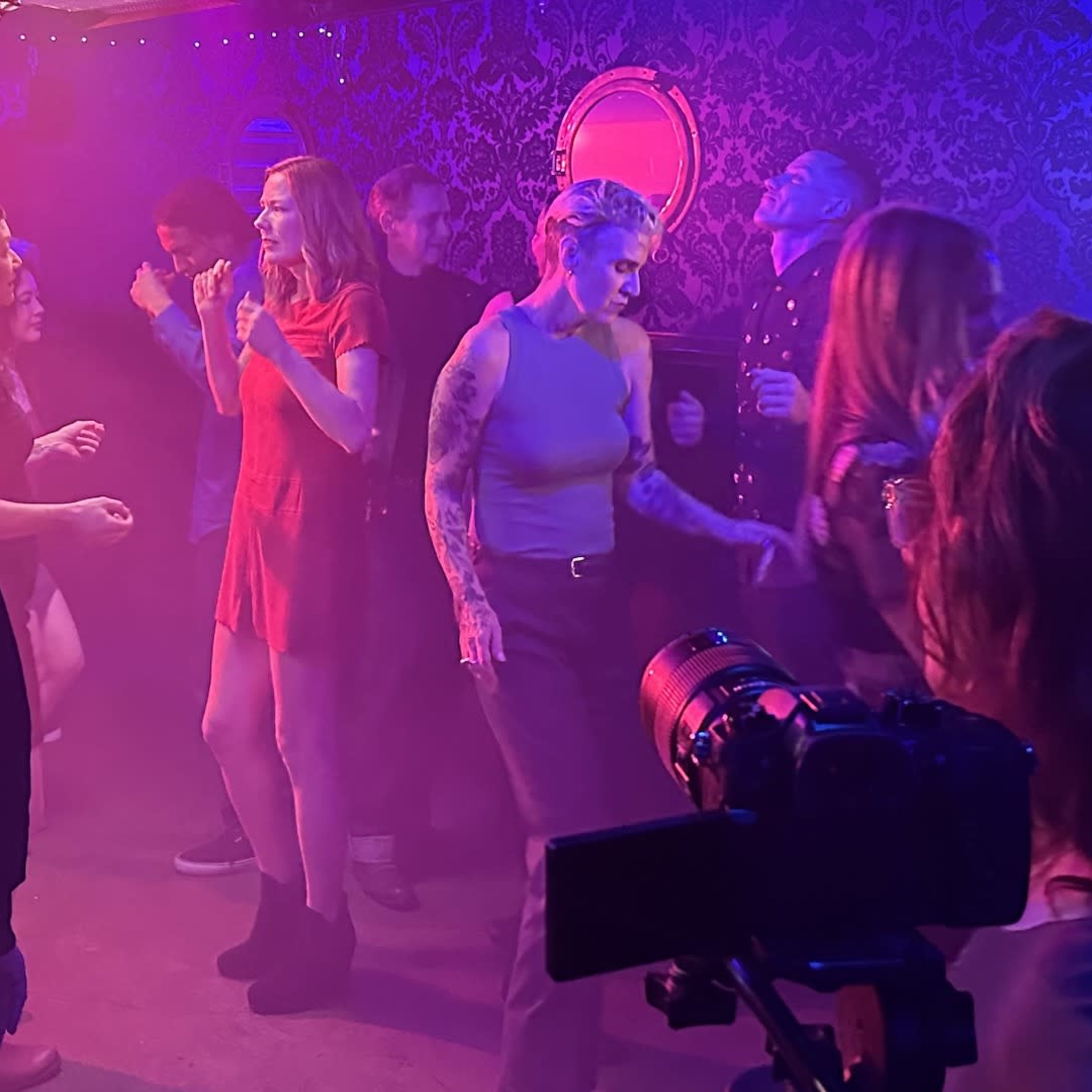 A group of people dance in a fog-filled, dimly lit room with colorful lights and decorative mirrors on the walls.