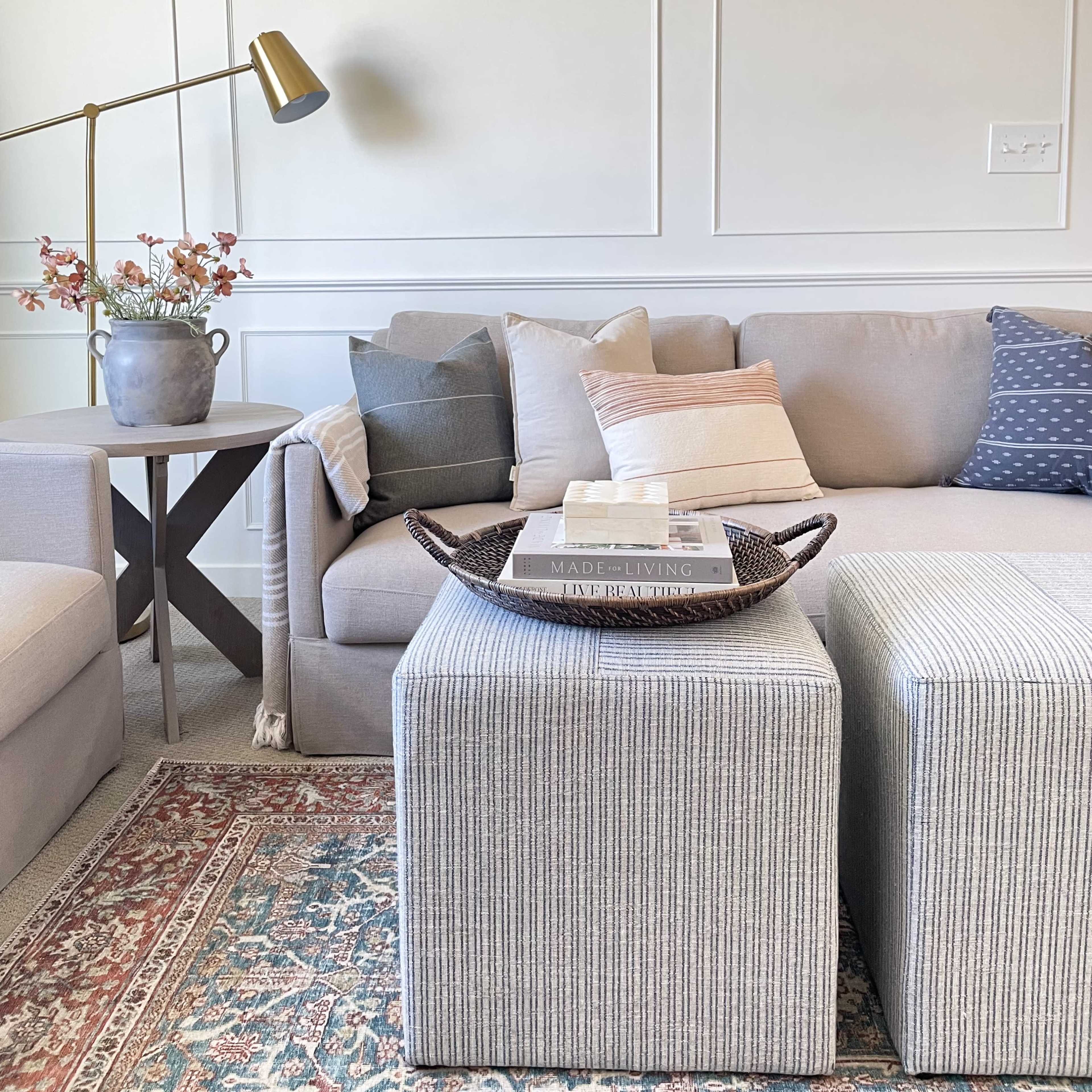 A light-colored living room features a gray sofa with various throw pillows, a woven tray holding books on a striped ottoman, and a floor lamp beside a vase of flowers.