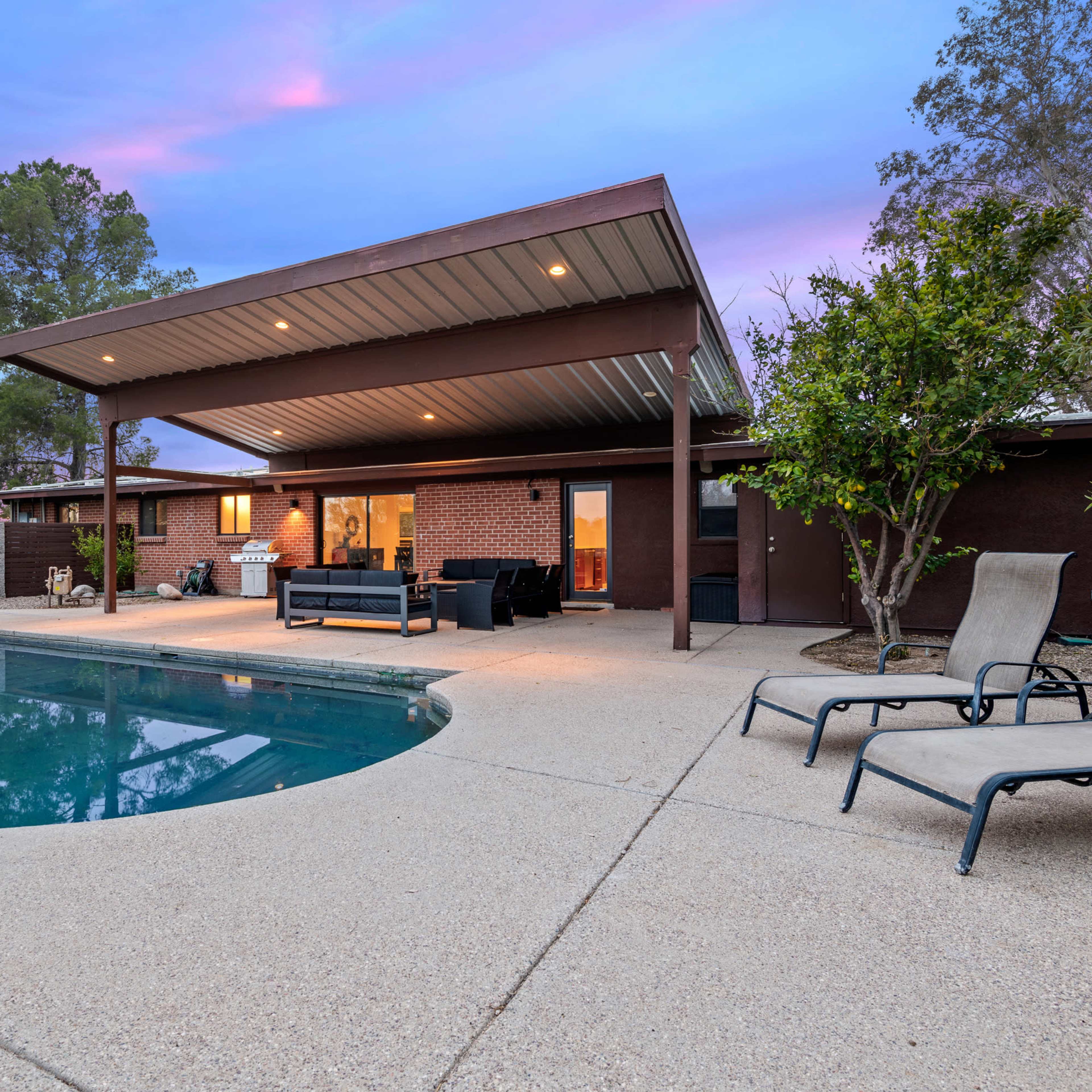 A backyard with a pool surrounded by patio furniture and a house with a covered area, set against a colorful sky at dusk.