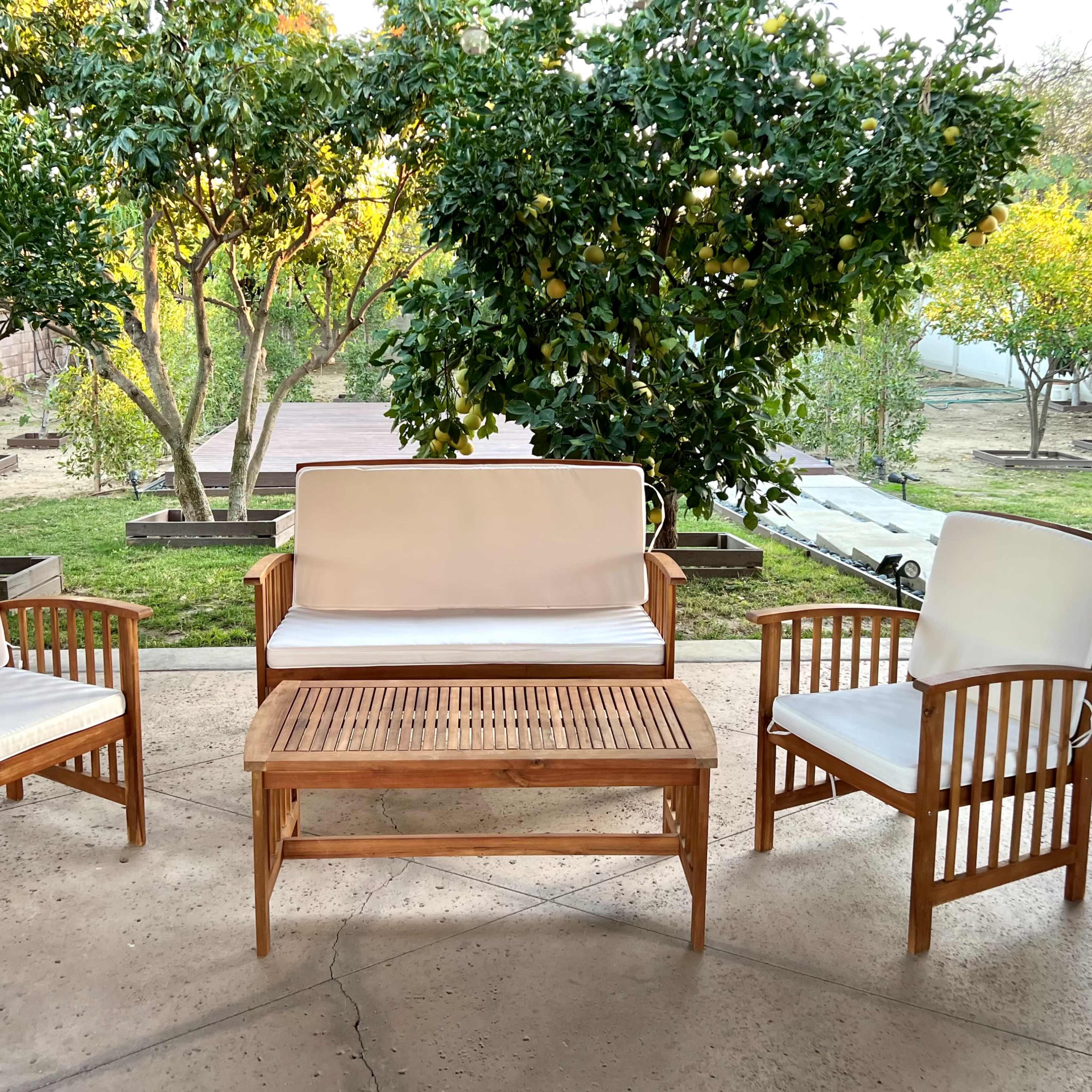 A set of wooden patio chairs and a table are arranged in a garden surrounded by trees.